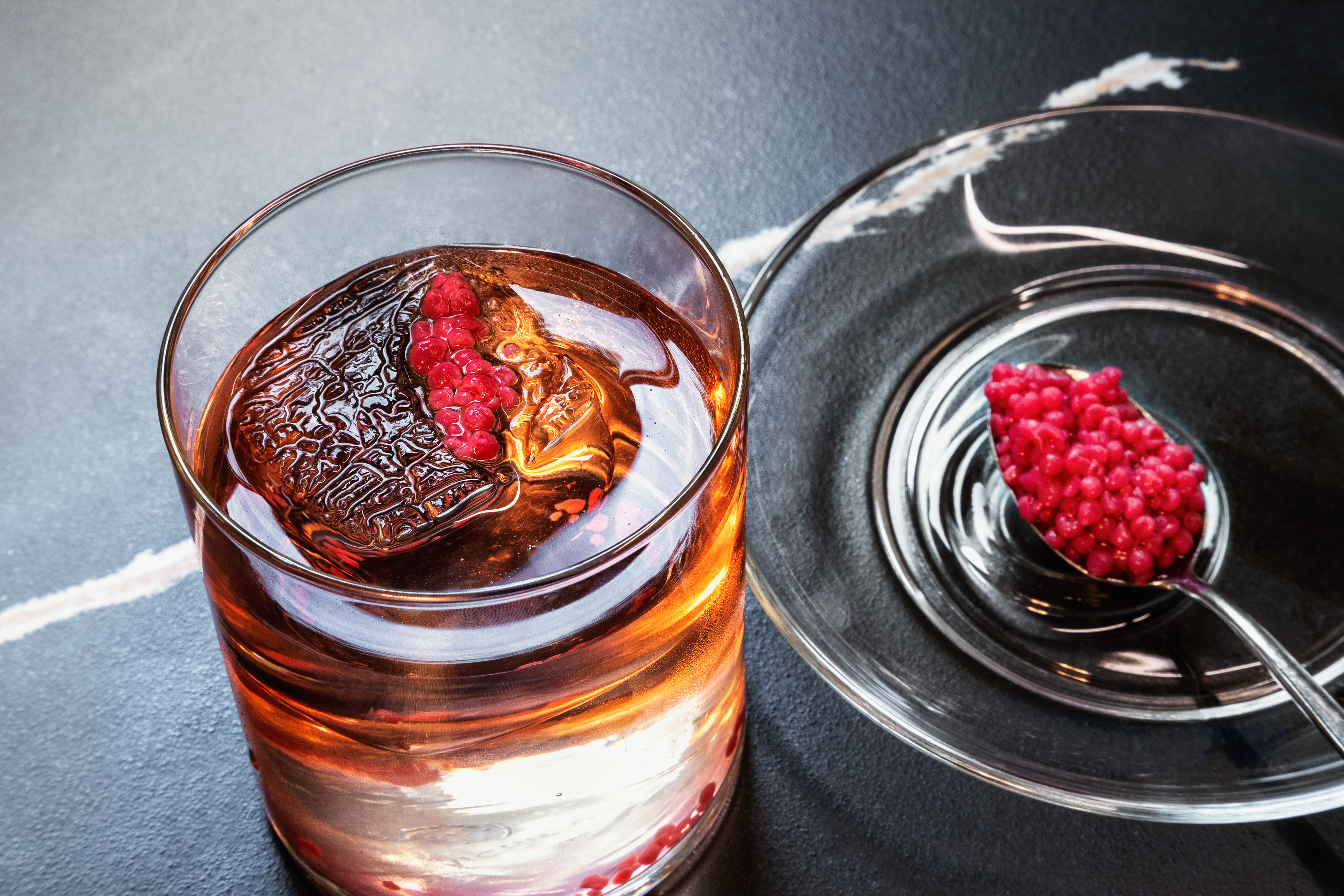 Elevated cocktail with raspberries, textured ice, and raspberry spheres on a spoon.