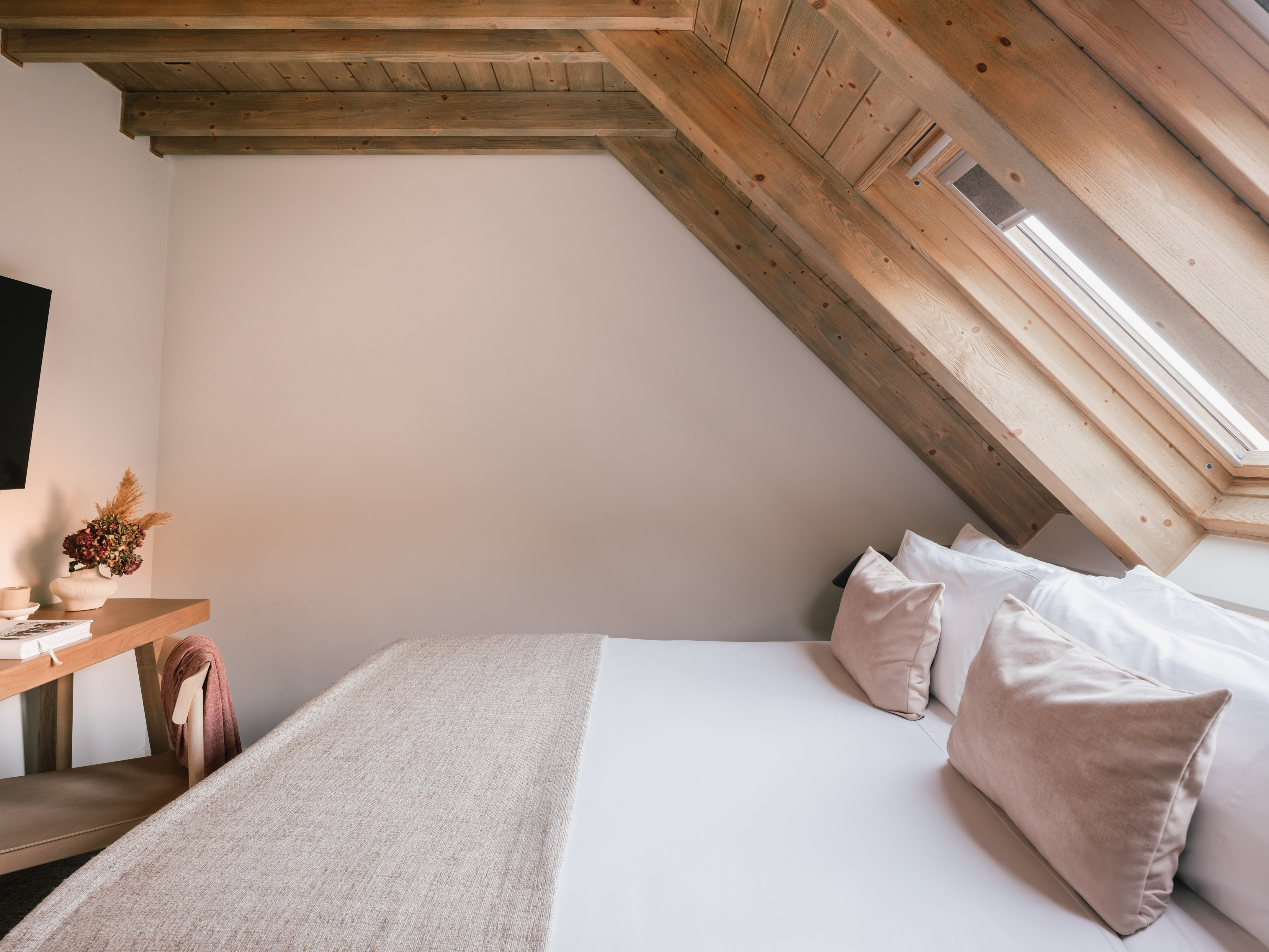 Cozy attic bedroom with wooden beams, white bed, skylight, and small desk.