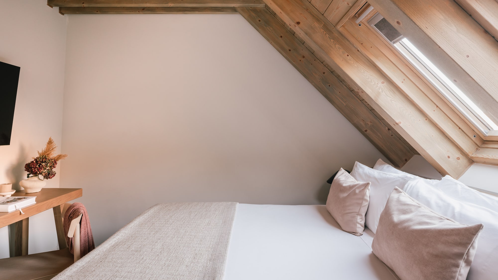 Cozy attic bedroom with wooden beams, white bed, skylight, and small desk.
