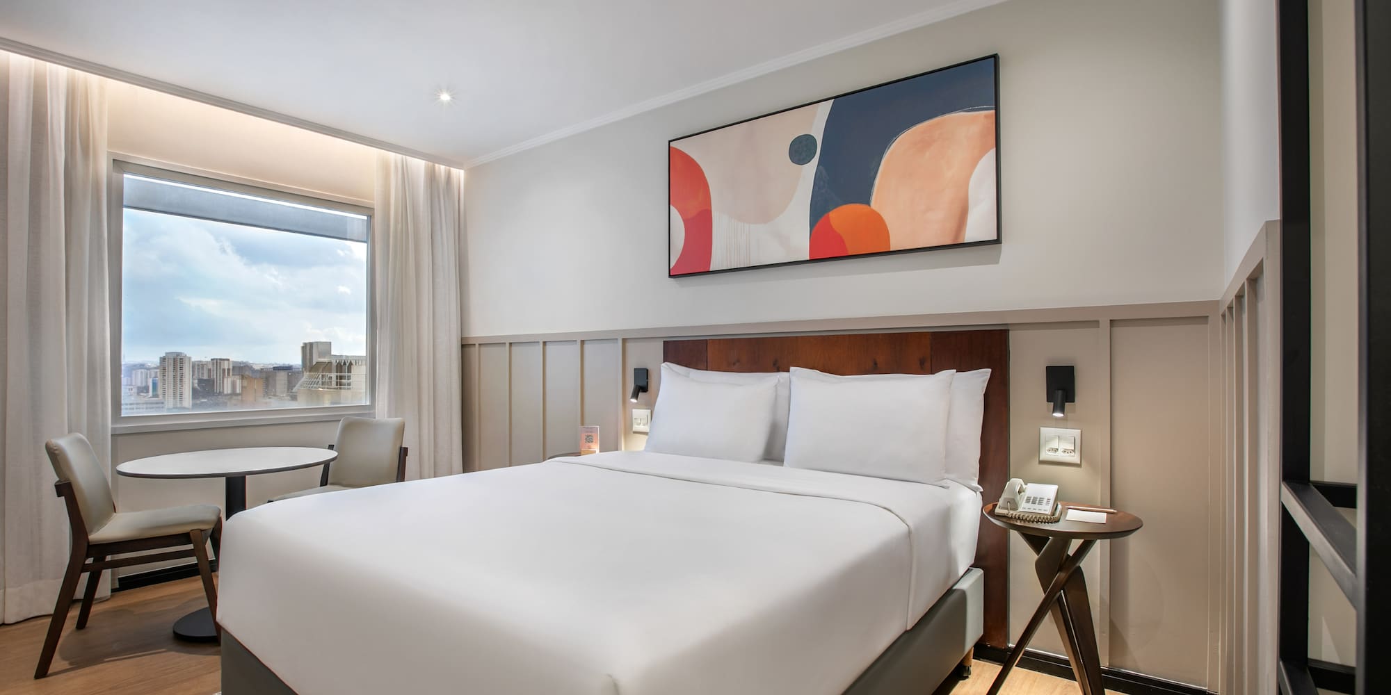 Modern hotel bedroom with king bed, abstract art, city view, and seating.