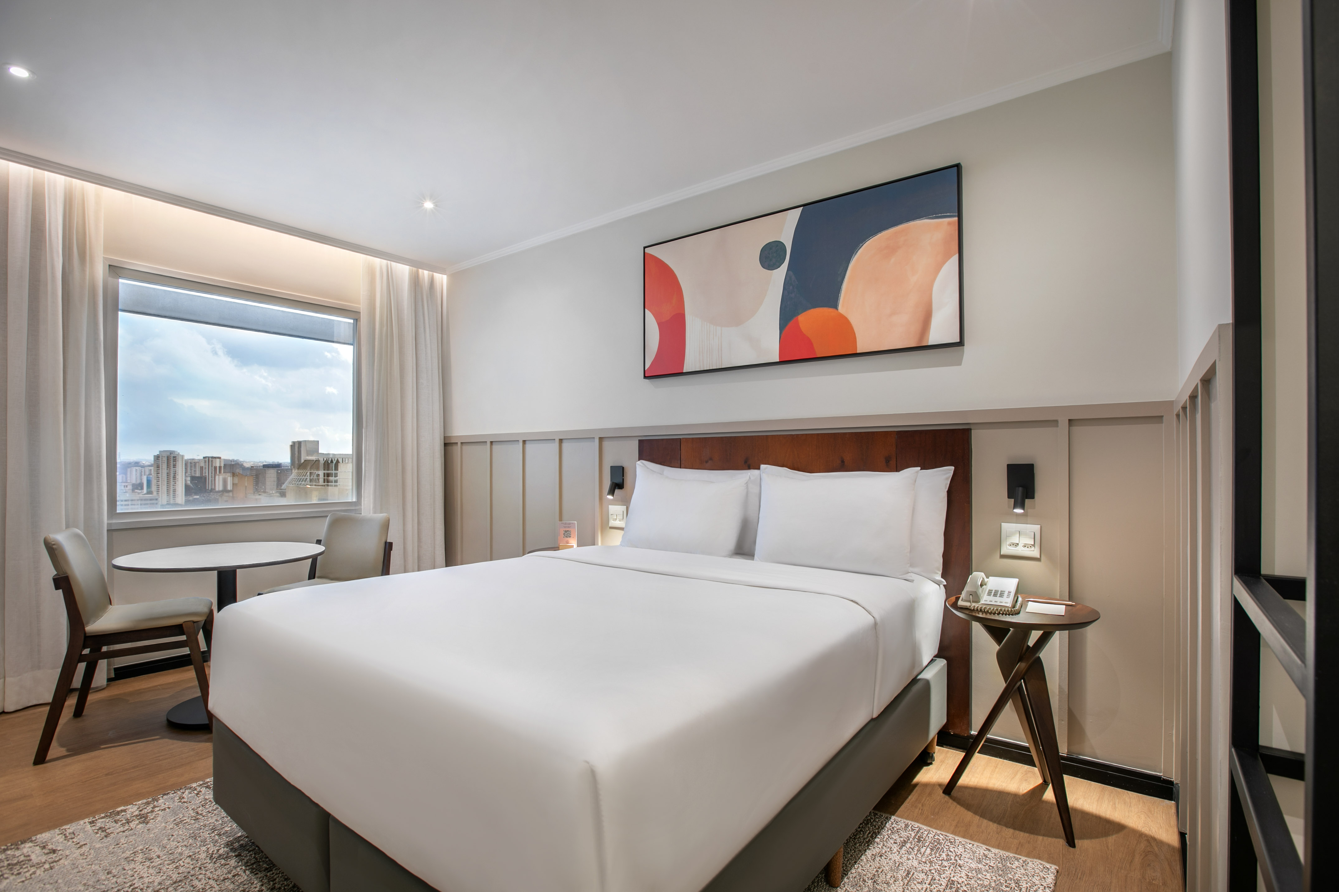 Modern hotel bedroom with king bed, abstract art, city view, and seating.