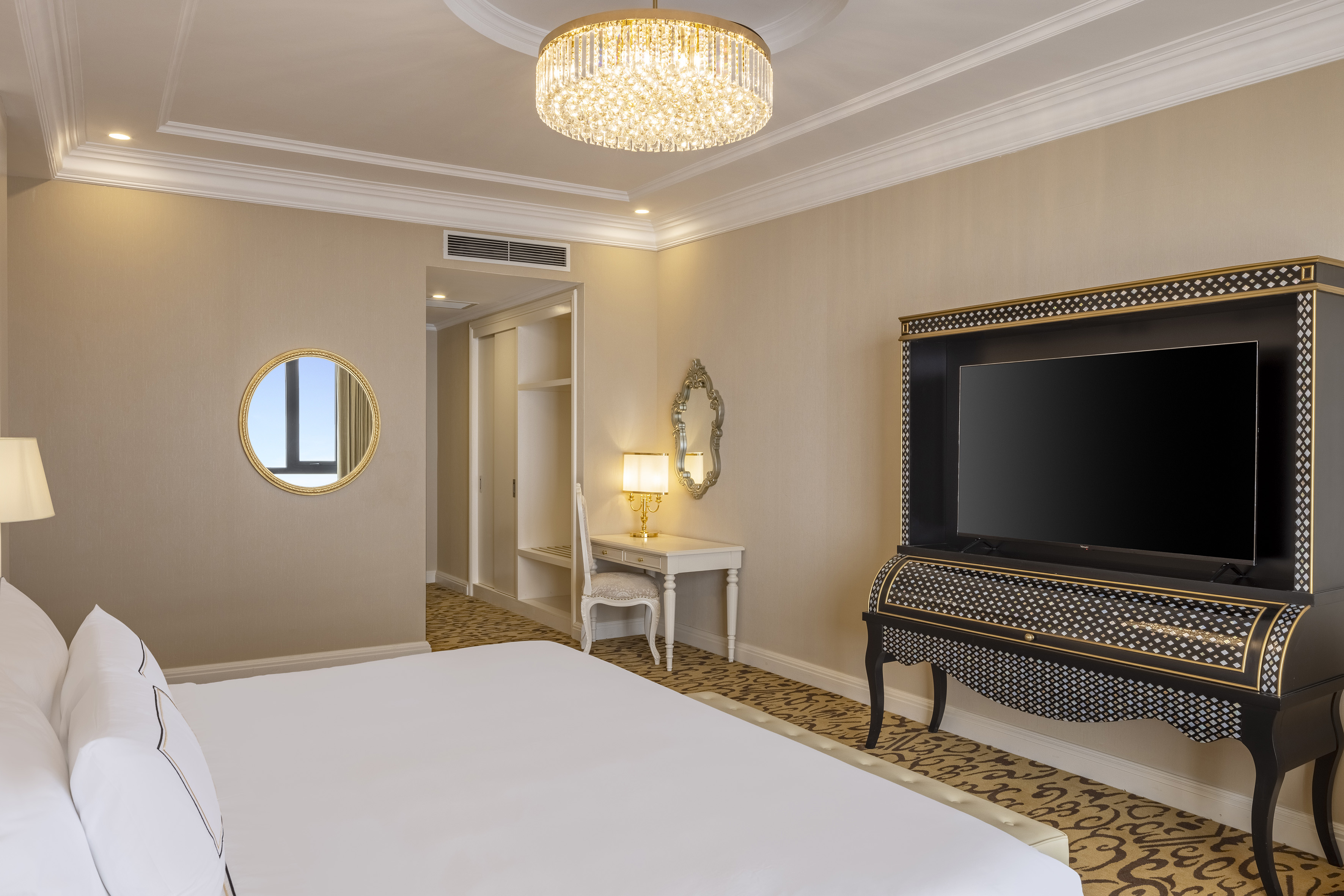 Elegant hotel suite with luxurious bed, ornate TV console, and crystal chandelier.