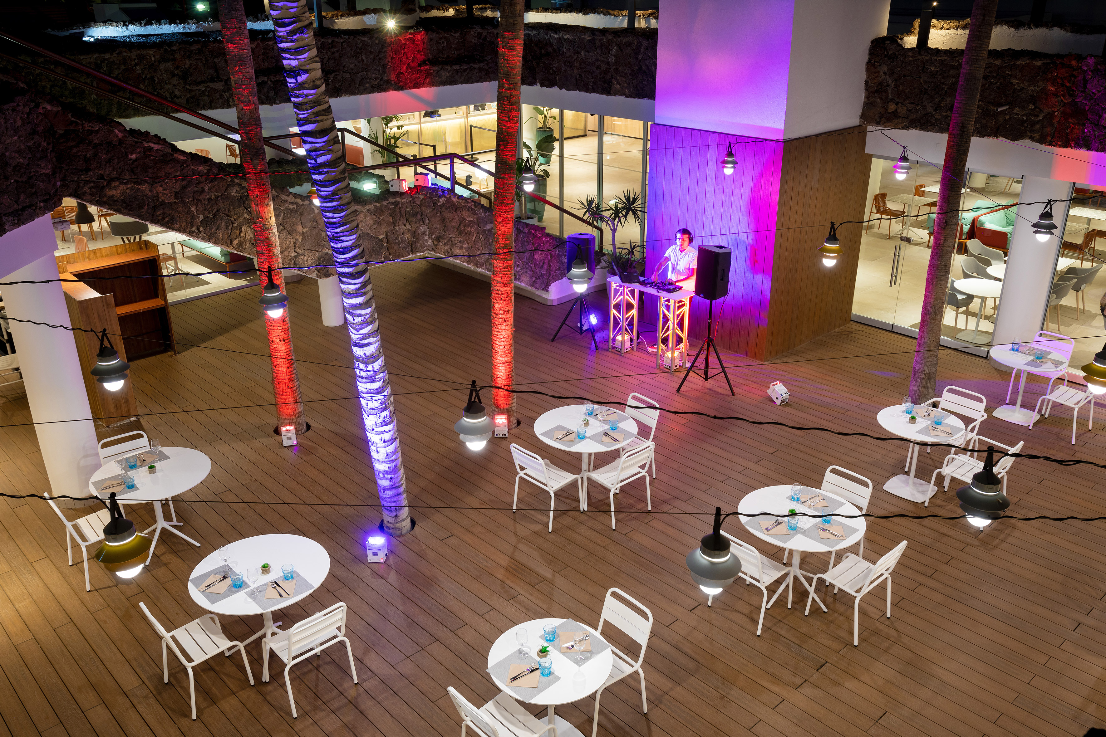 Evening resort patio with DJ, palm trees lit purple and red, and dining tables.