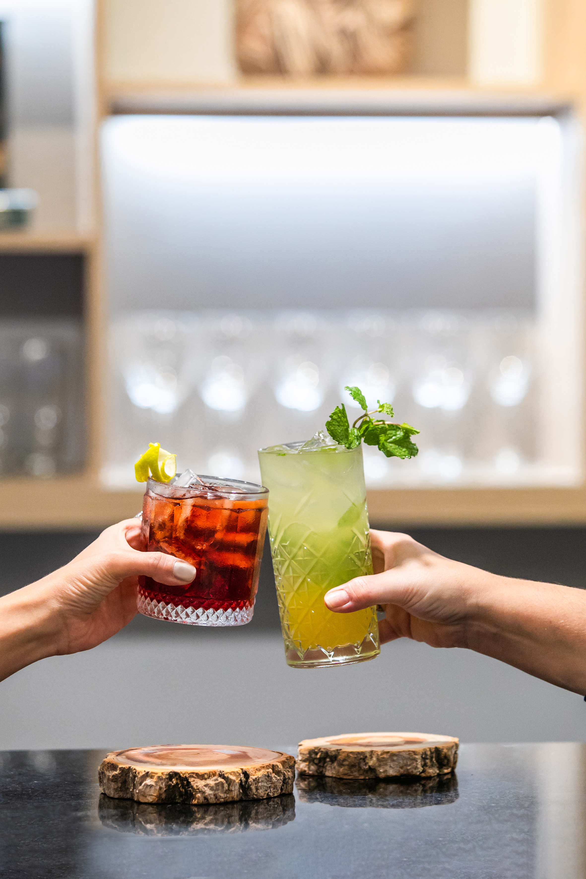 Hands toasting with a red Negroni and green mint cocktail at a bar.