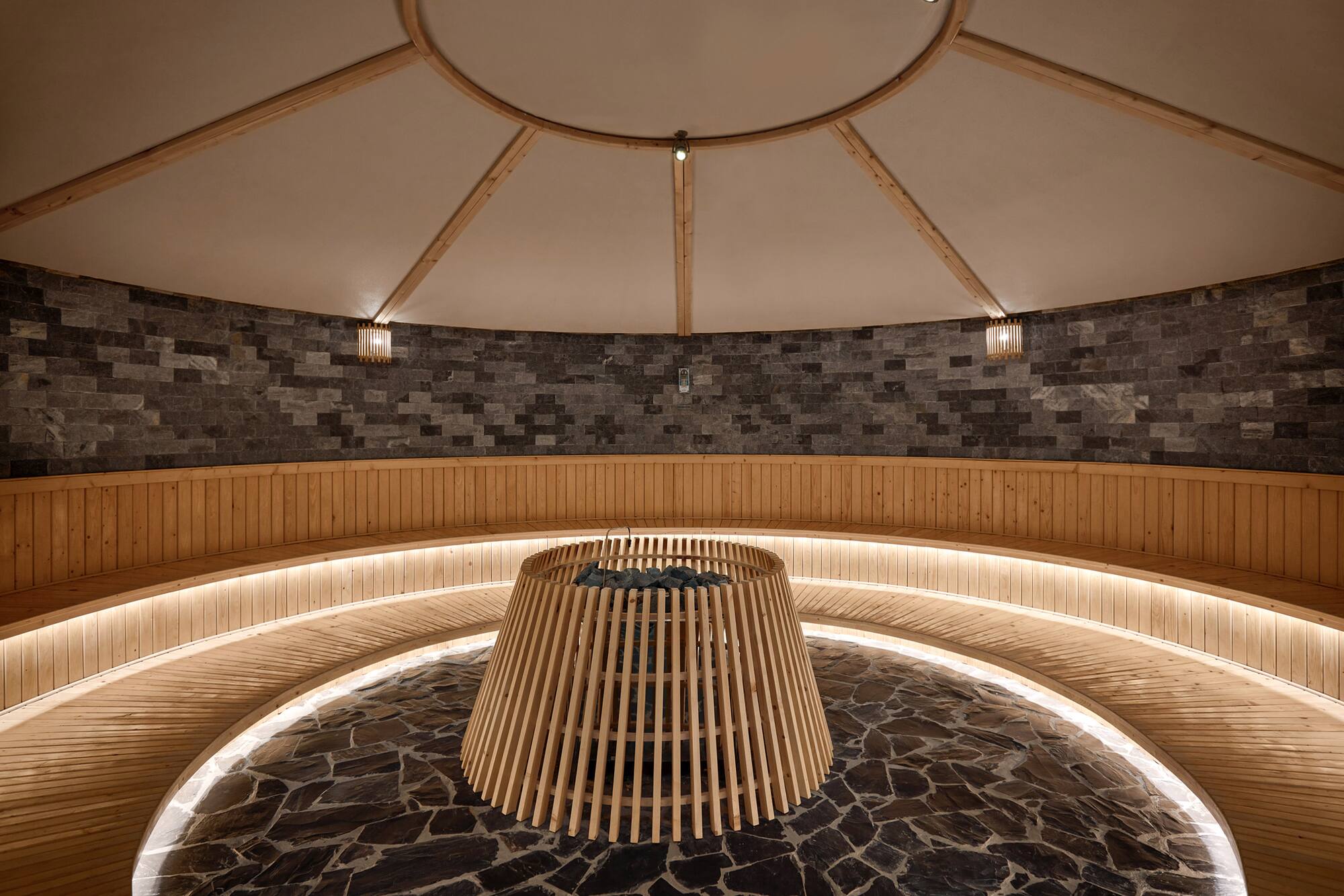 Circular sauna with stone walls, curved wooden benches, and central heater.