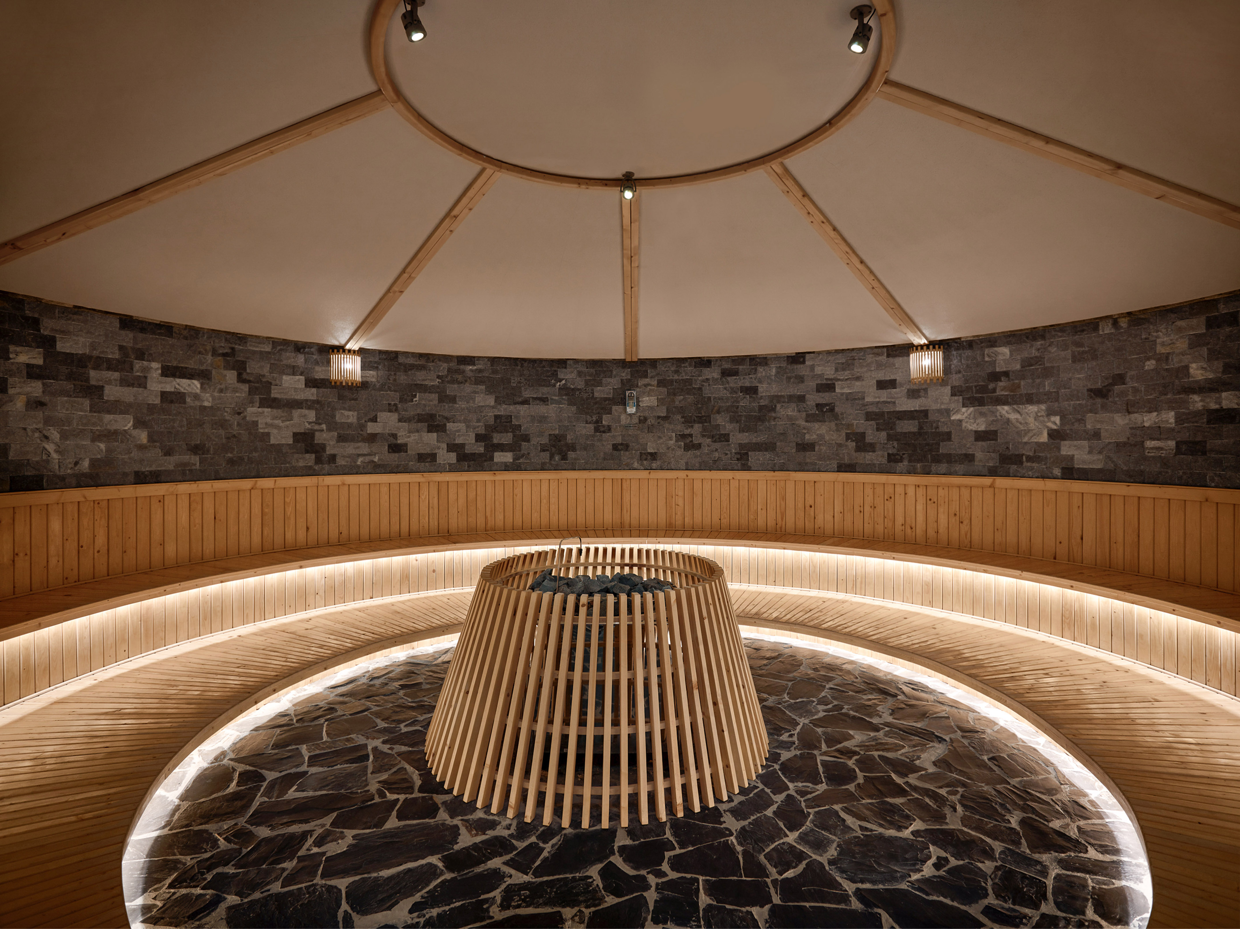 Circular sauna with stone walls, curved wooden benches, and central heater.