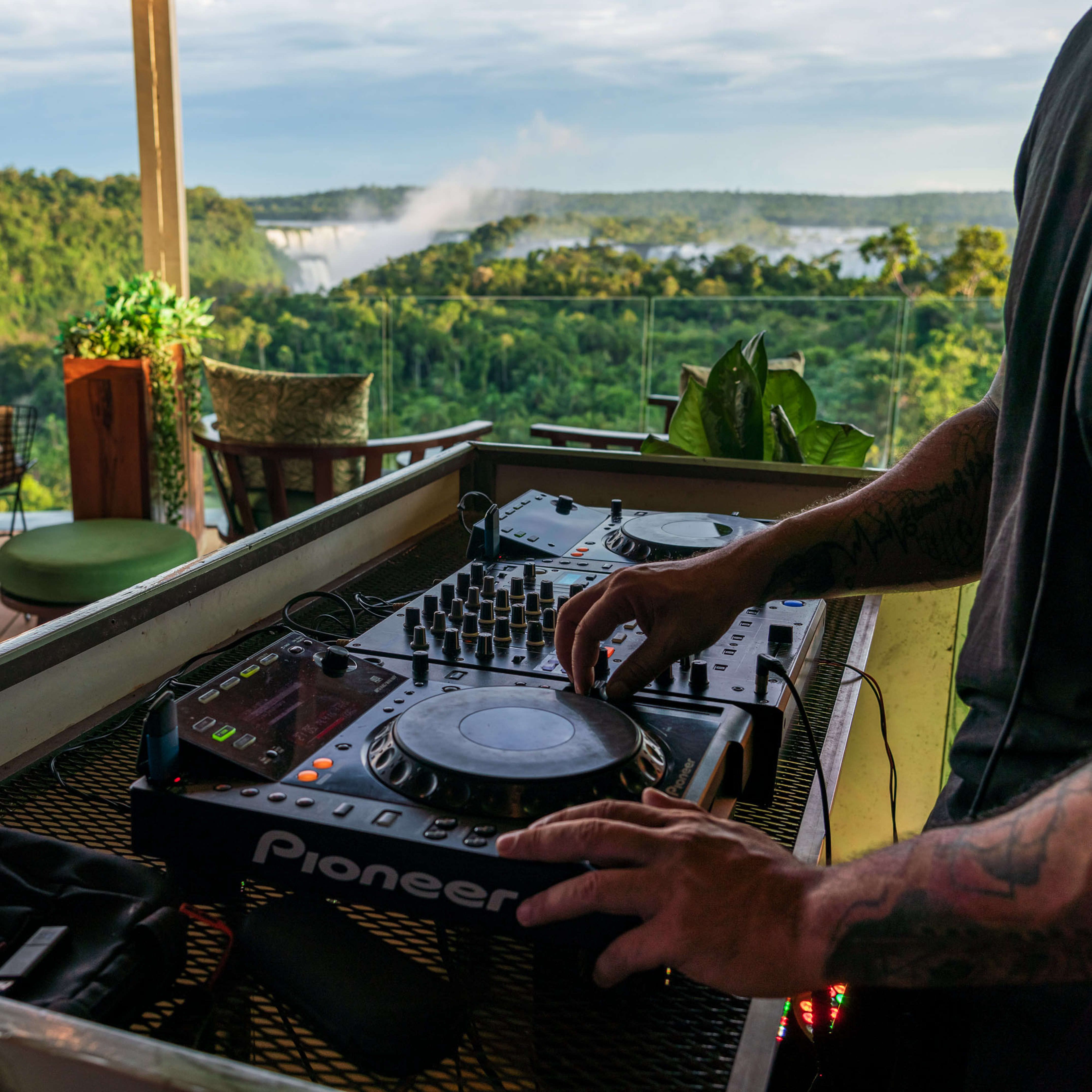 DJ's hands on Pioneer mixer, overlooking Iguazu Falls and lush rainforest.