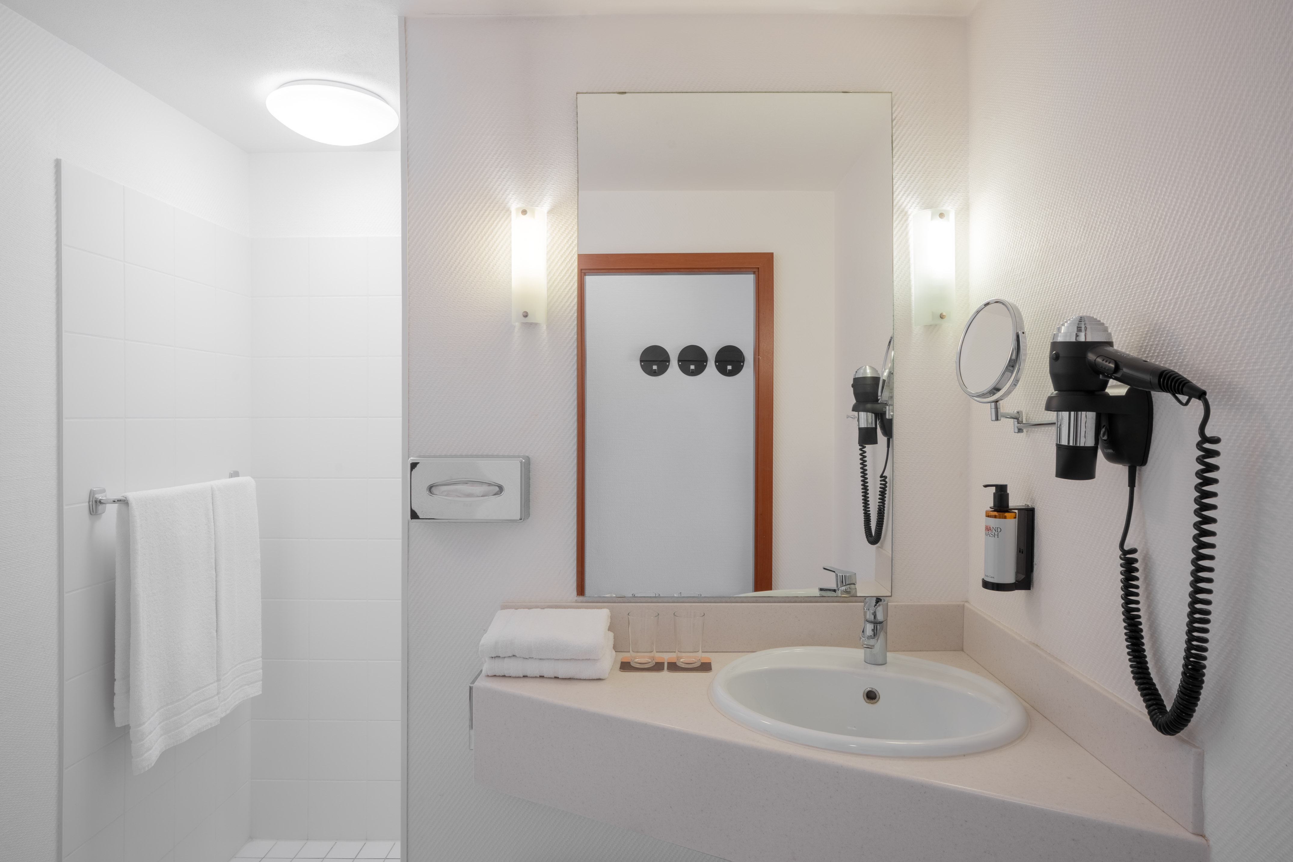 Clean white hotel bathroom with sink, mirror, shower, and hairdryer.