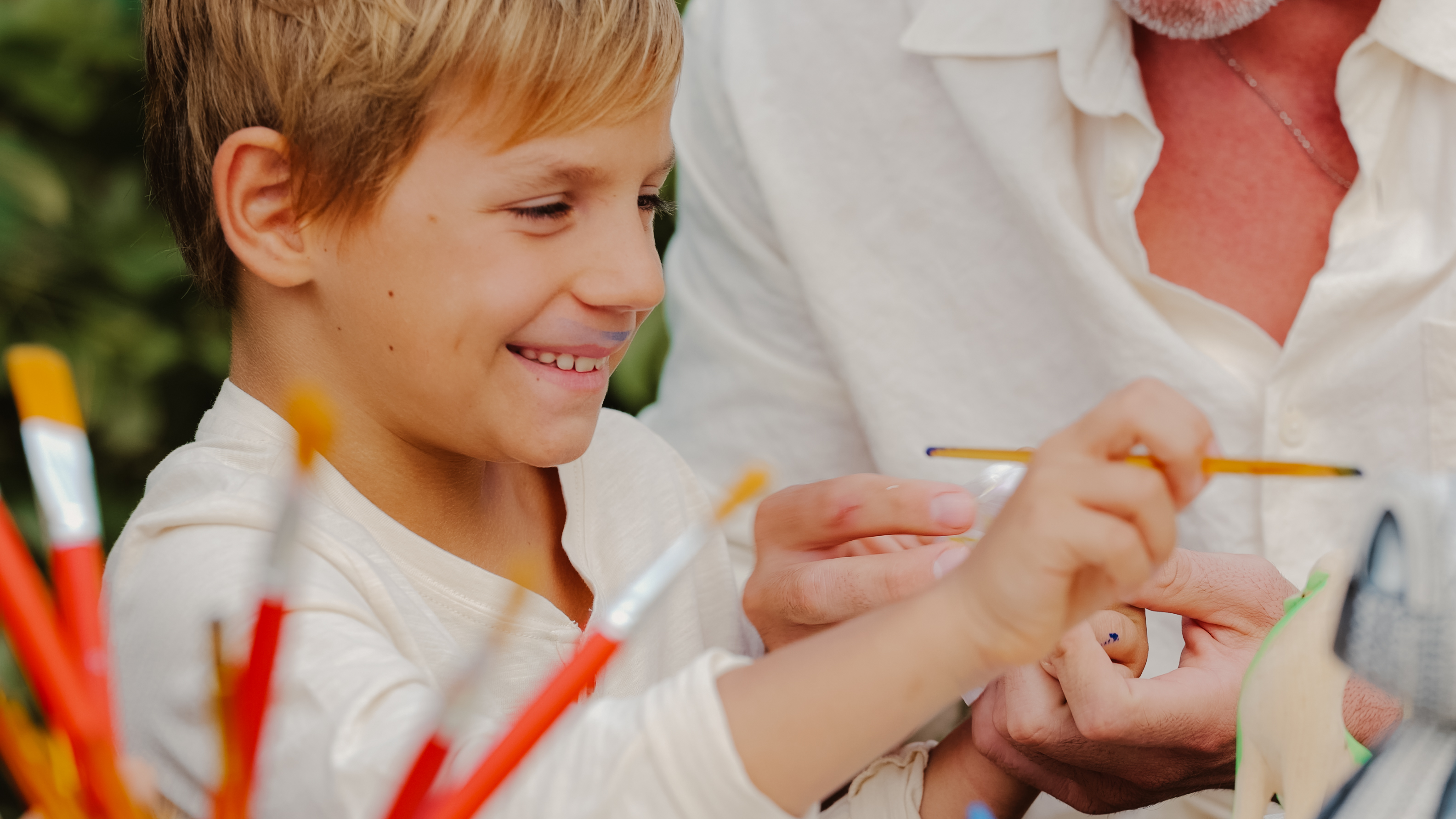 Boy with paint on lip smiles while painting with an adult.
