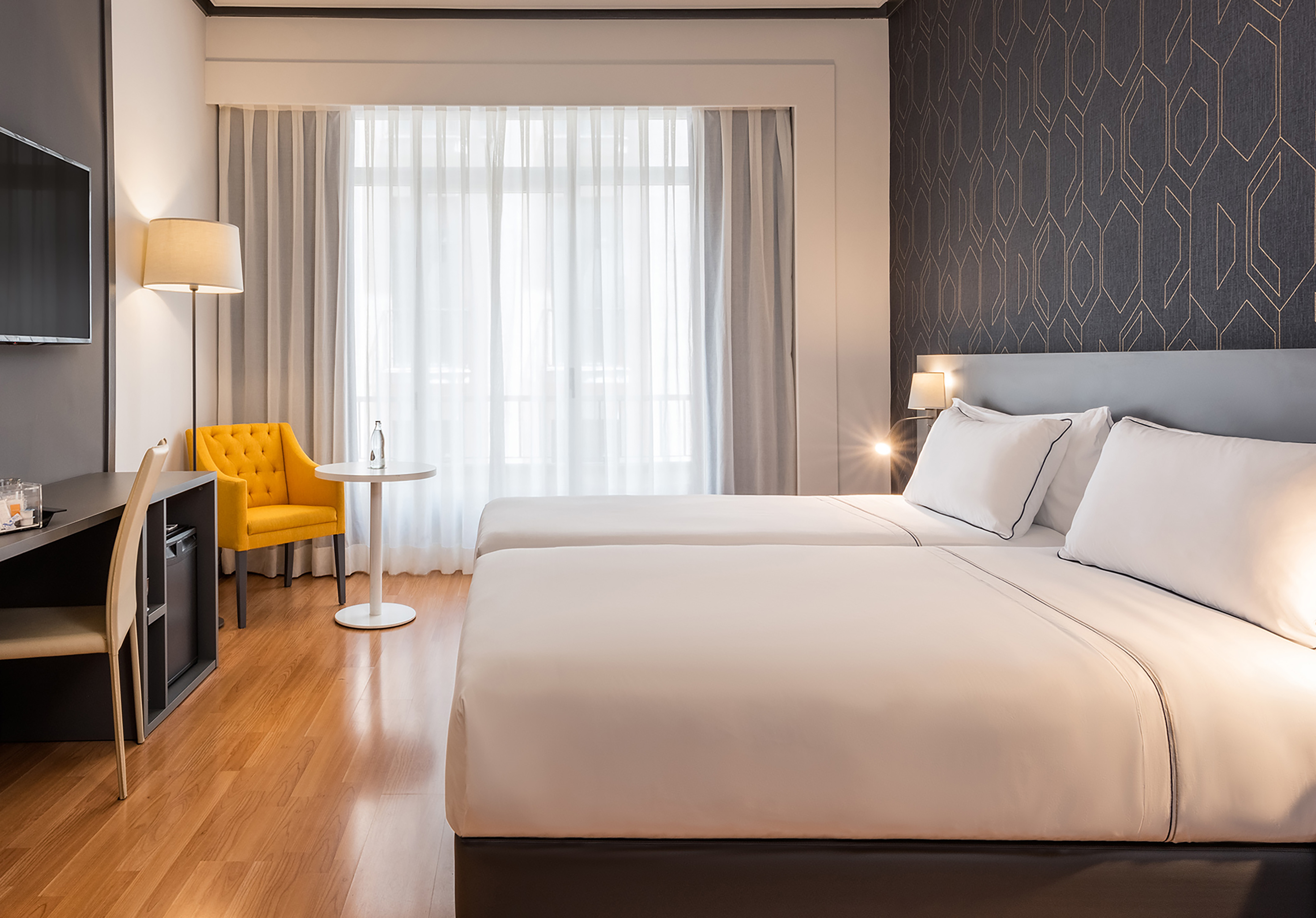 Modern hotel room with two single beds, yellow armchair, desk, and large window.