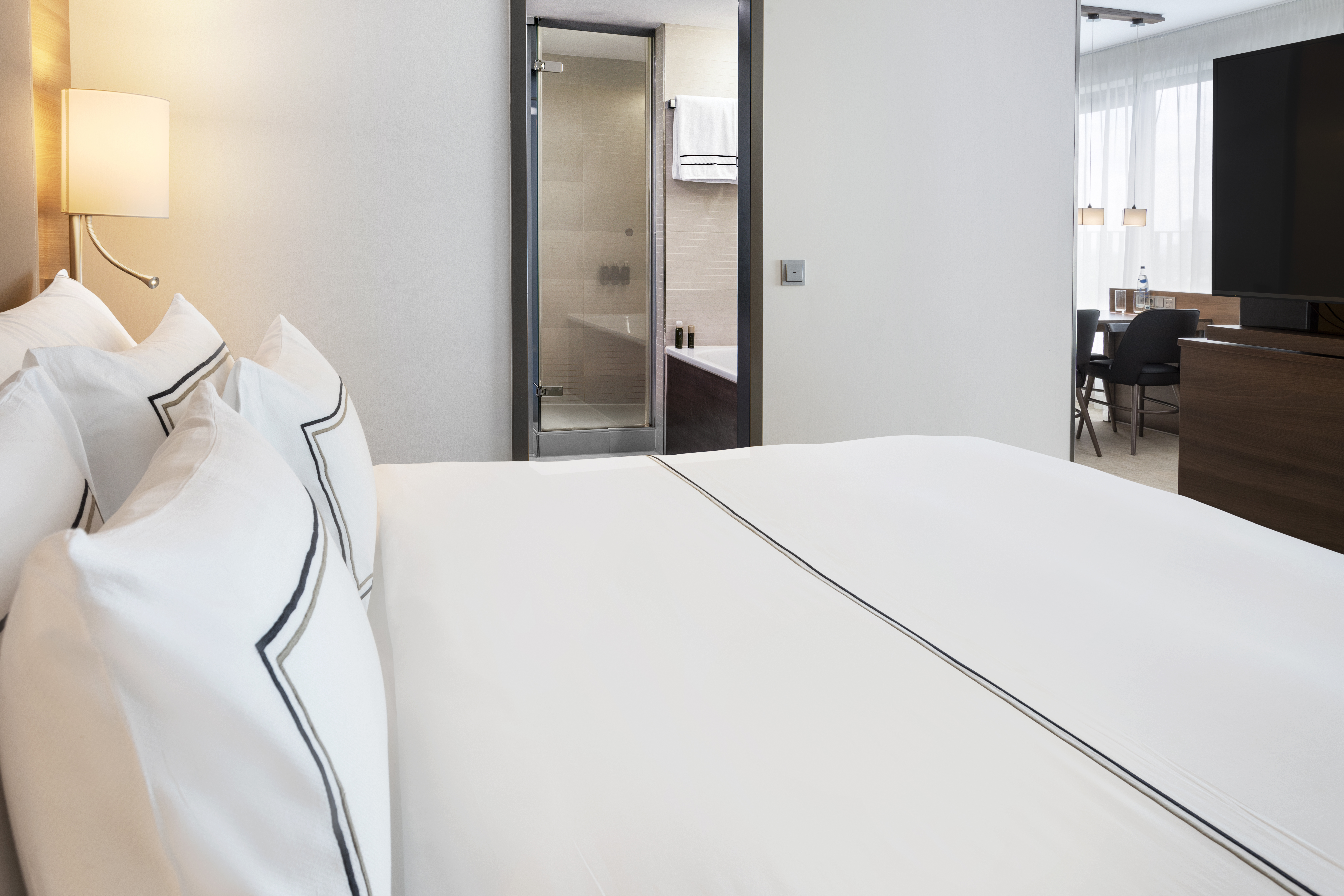 Contemporary hotel suite bedroom with white bed, ensuite bath, and sitting area.