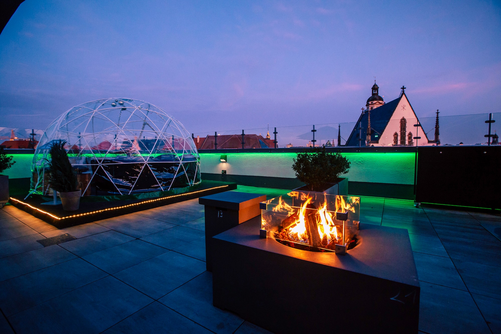 Evening rooftop terrace with igloo, glowing fire pit, green lights, city view.