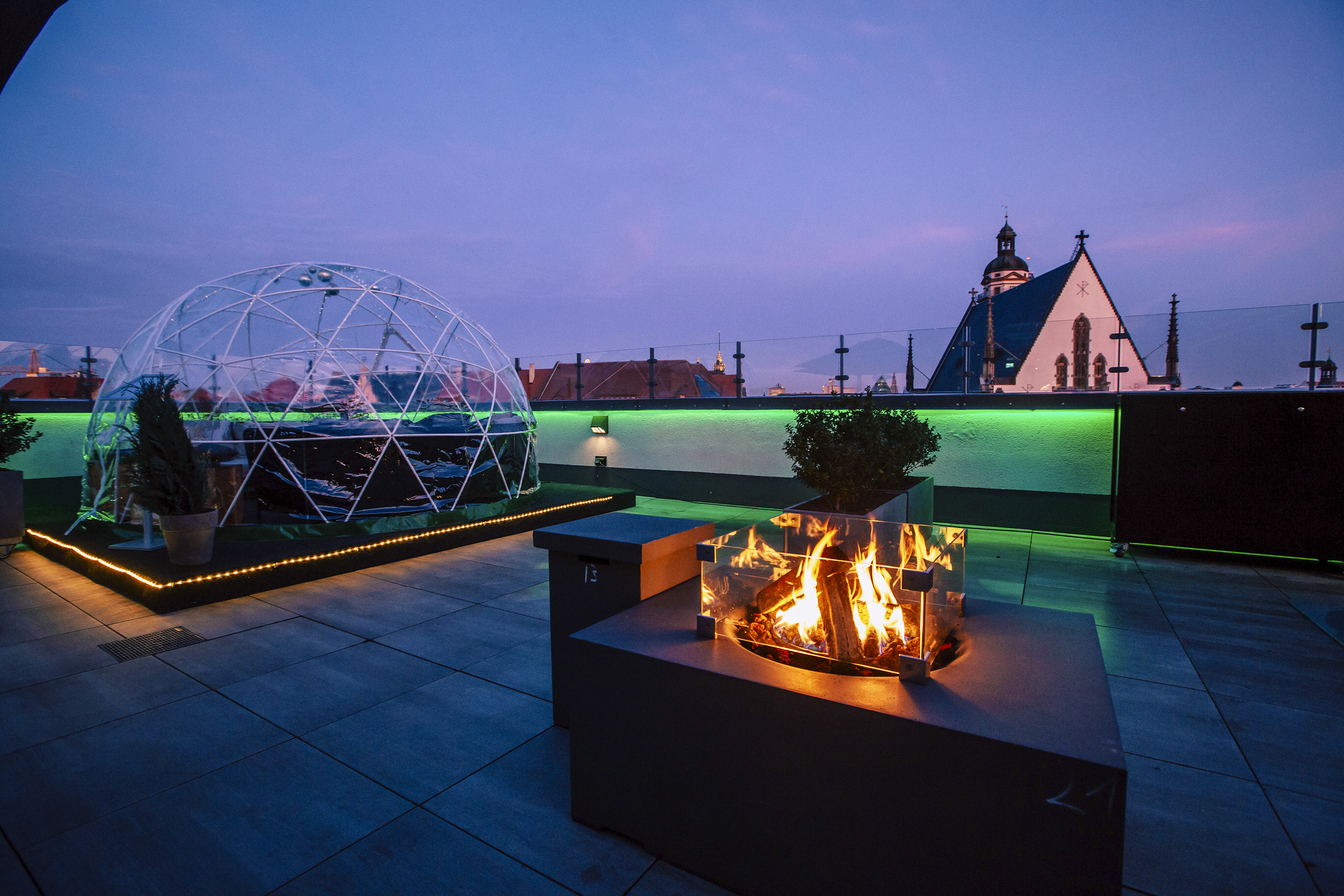 Evening rooftop terrace with igloo, glowing fire pit, green lights, city view.