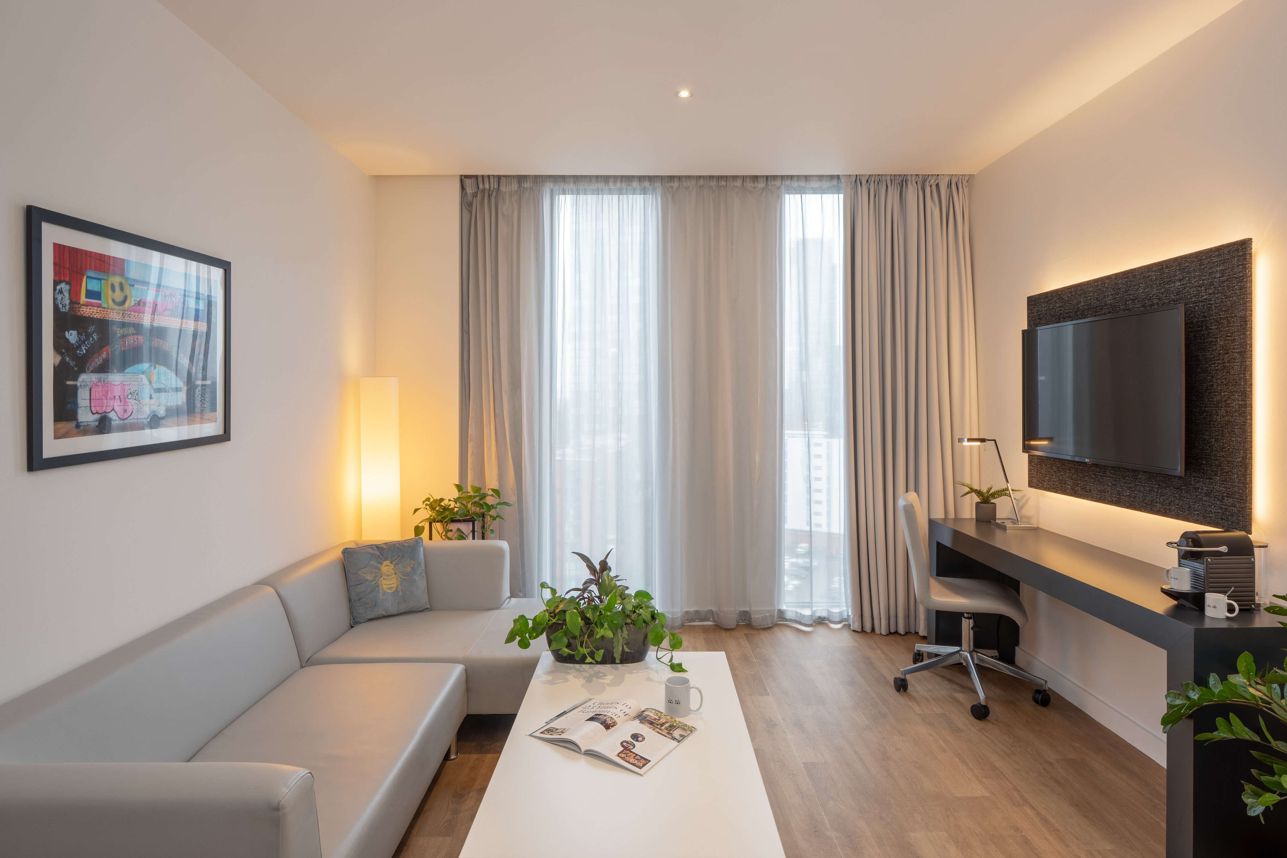 Modern hotel suite living room with grey sofa, work desk, mounted TV, and window.