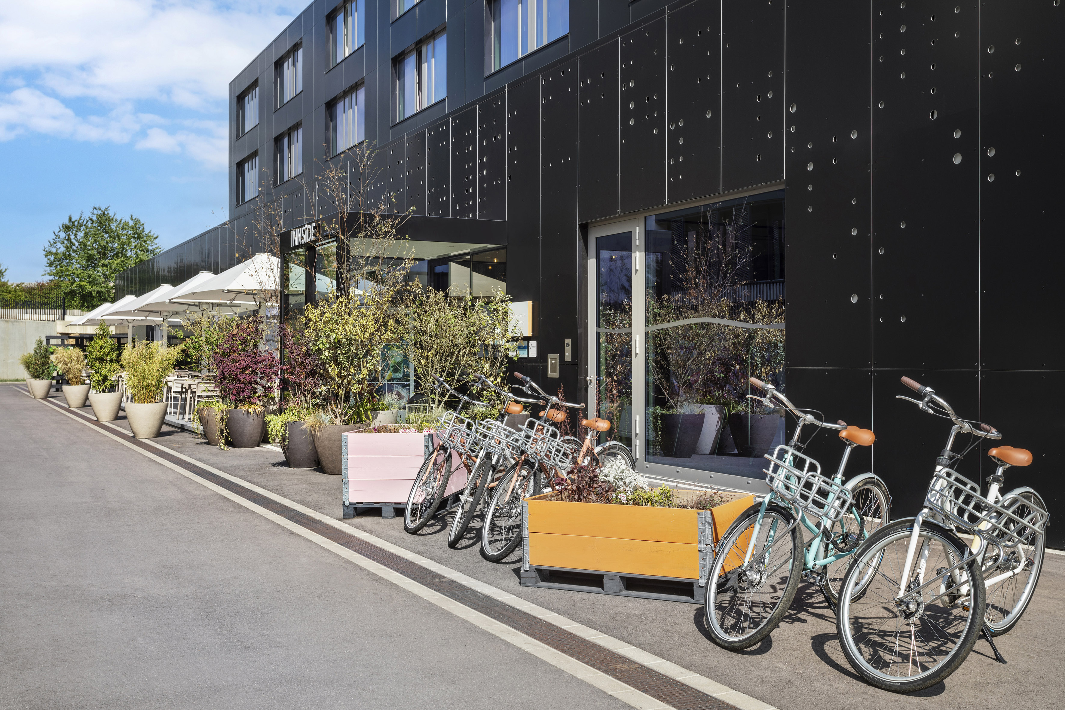 Black INNSIDE hotel facade with parked bicycles, plant beds, and outdoor seating. - INNSiDE Luxembourg, Люксембург