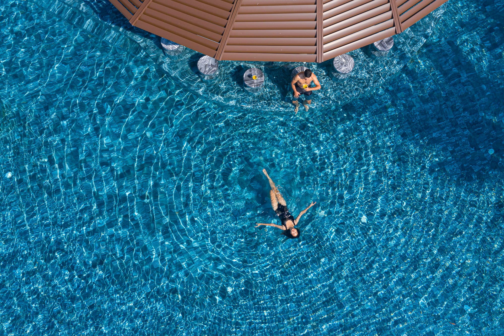 Aerial view of man at swim-up bar and woman floating in resort pool.