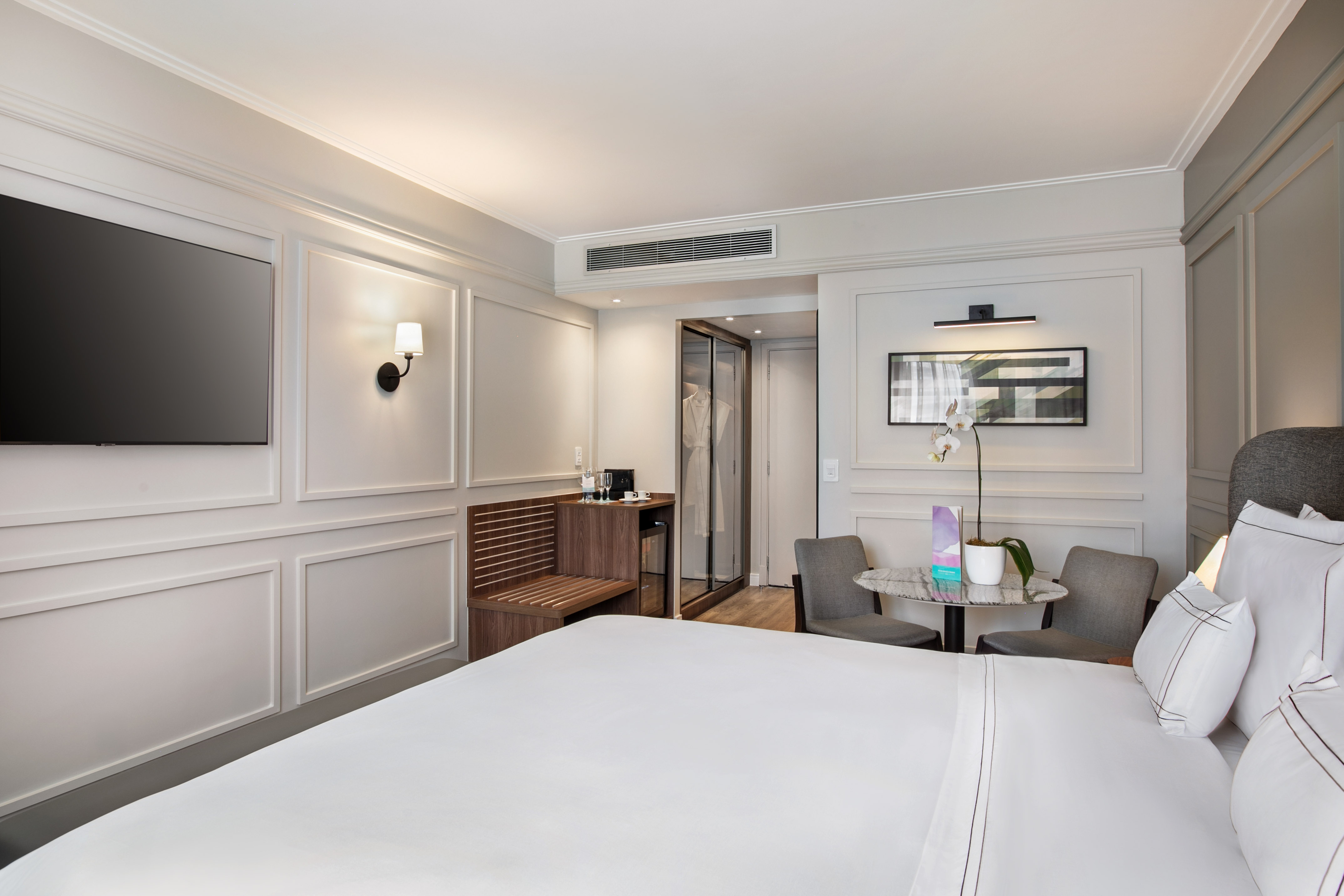 Elegant hotel room with large white bed, wall TV, wardrobe, and small seating area.