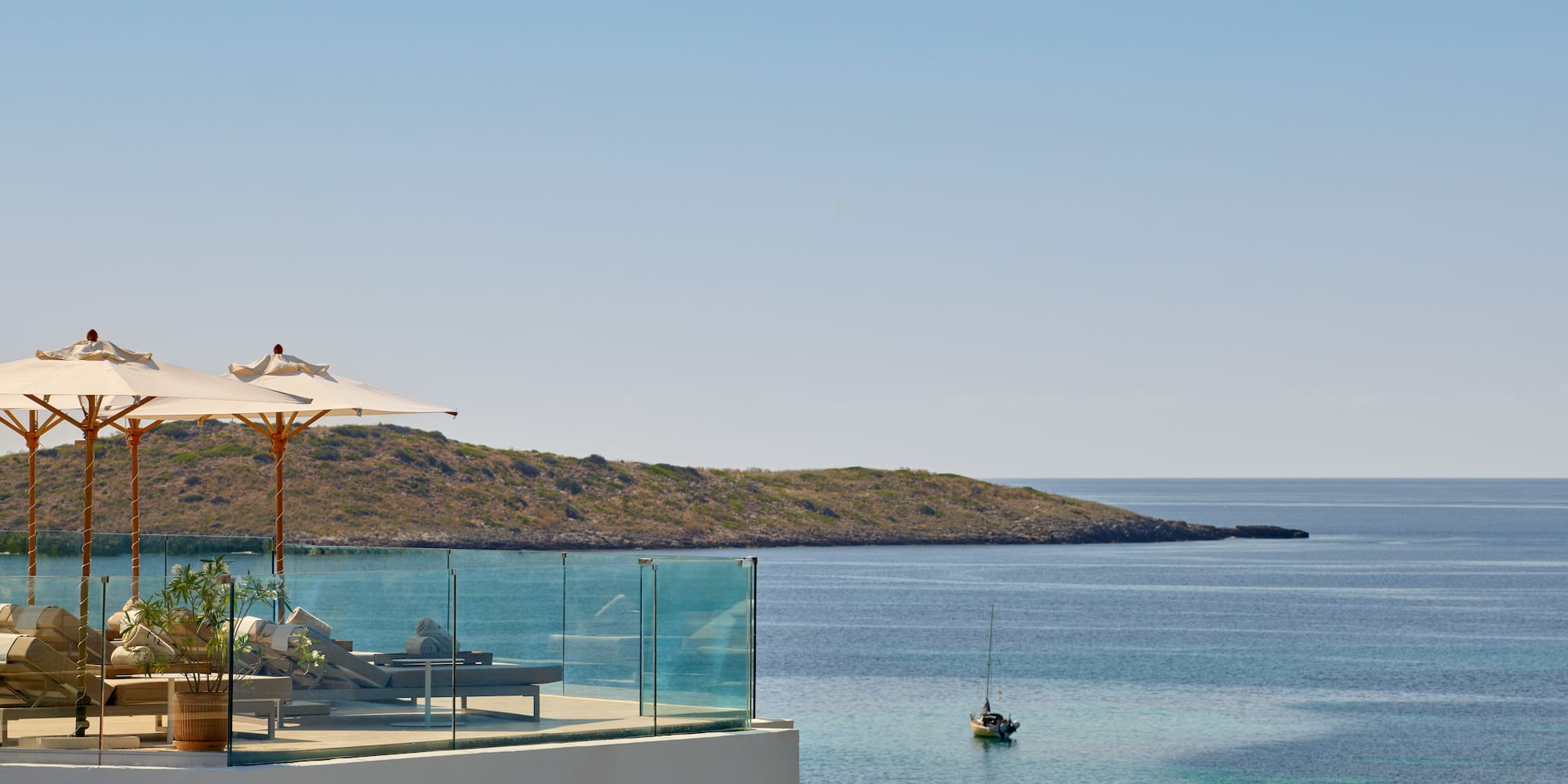 Terrace with sun loungers, umbrellas, overlooking calm bay, sailboat, island.