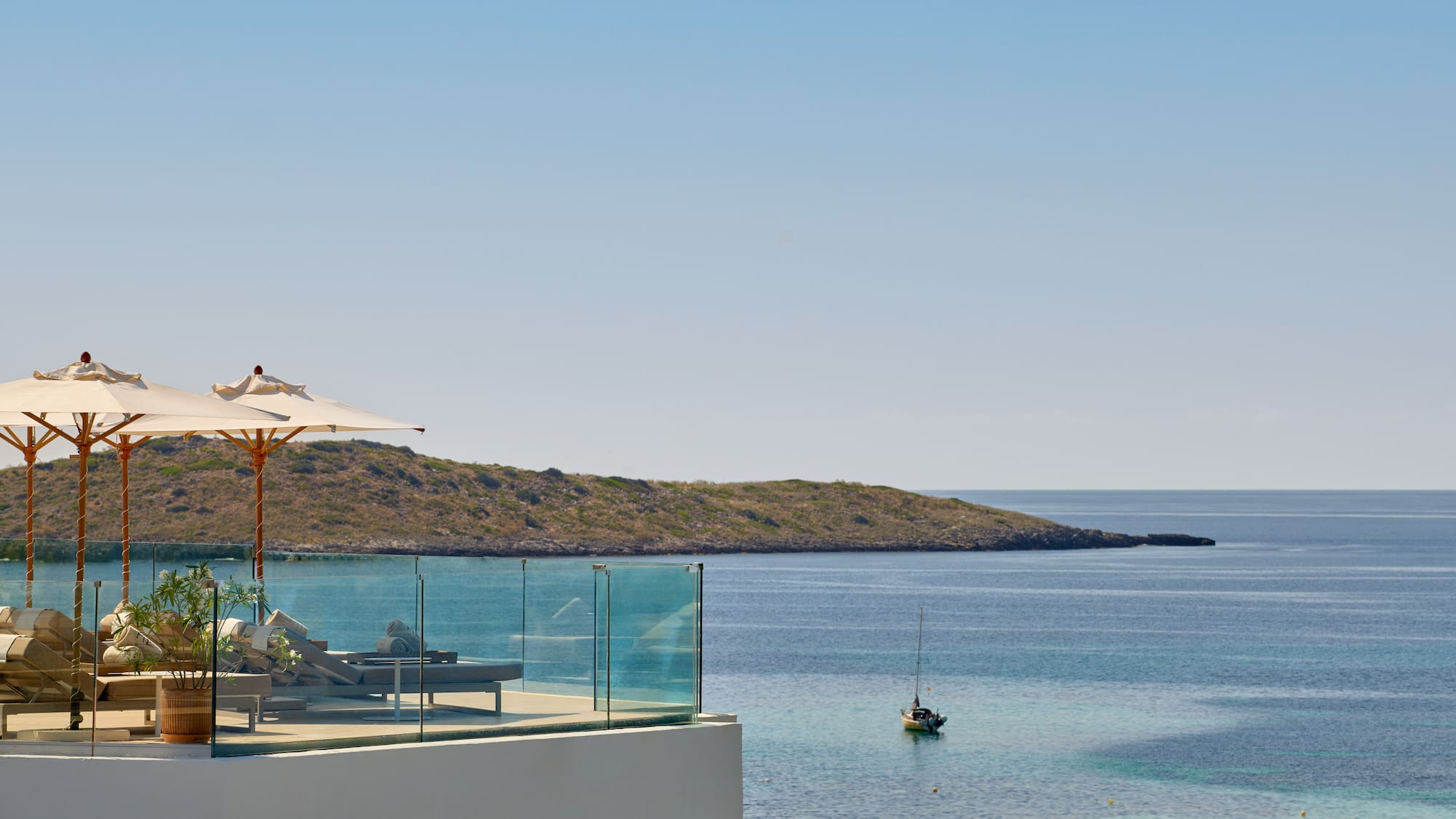 Terrace with sun loungers, umbrellas, overlooking calm bay, sailboat, island.