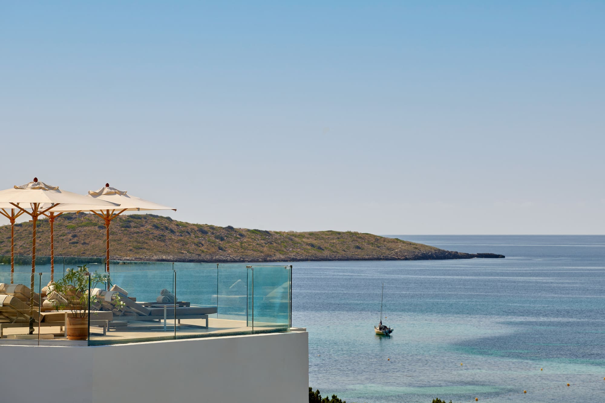 Terrace with sun loungers, umbrellas, overlooking calm bay, sailboat, island.