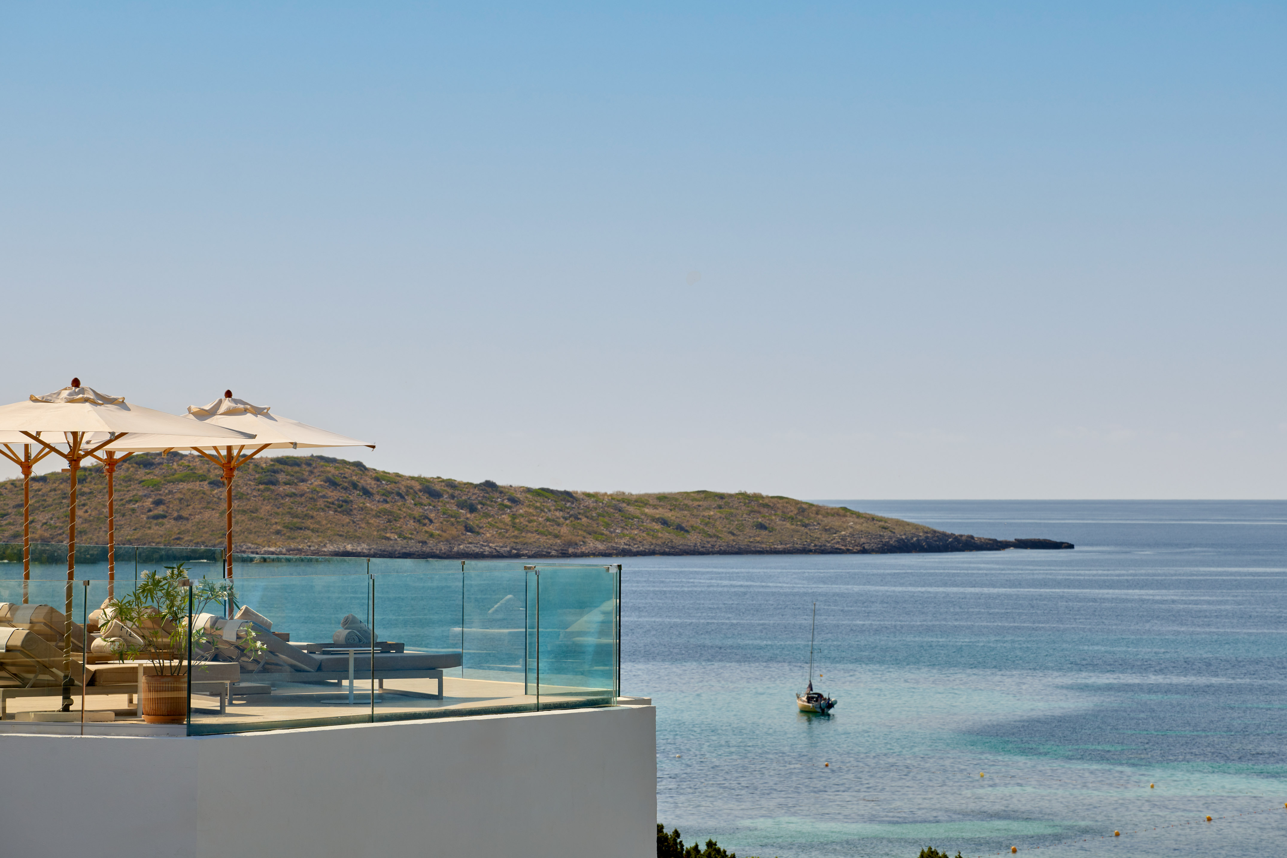 Terrace with sun loungers, umbrellas, overlooking calm bay, sailboat, island.