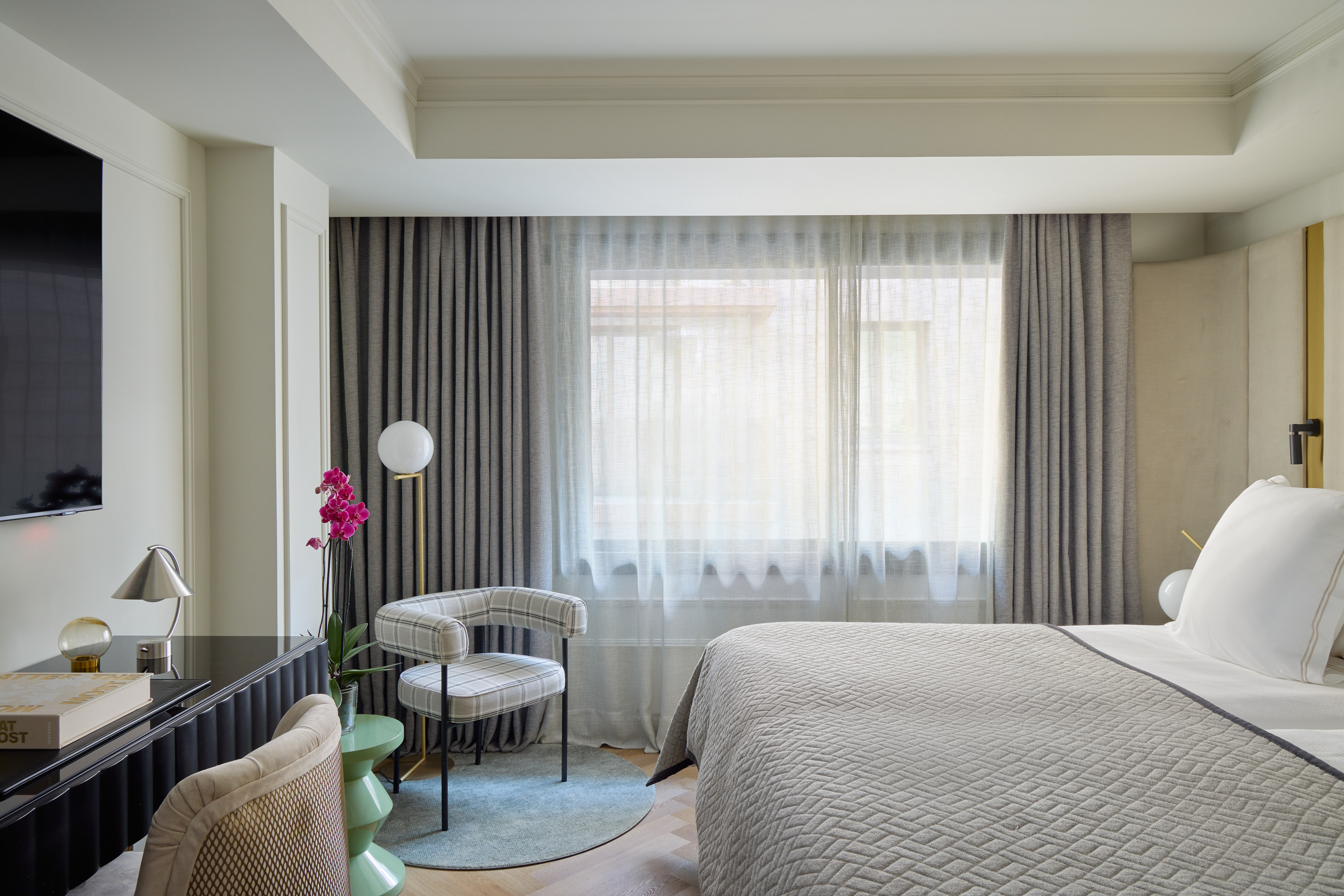 Modern hotel bedroom with grey bed, plaid armchair, sheer curtains, and desk.