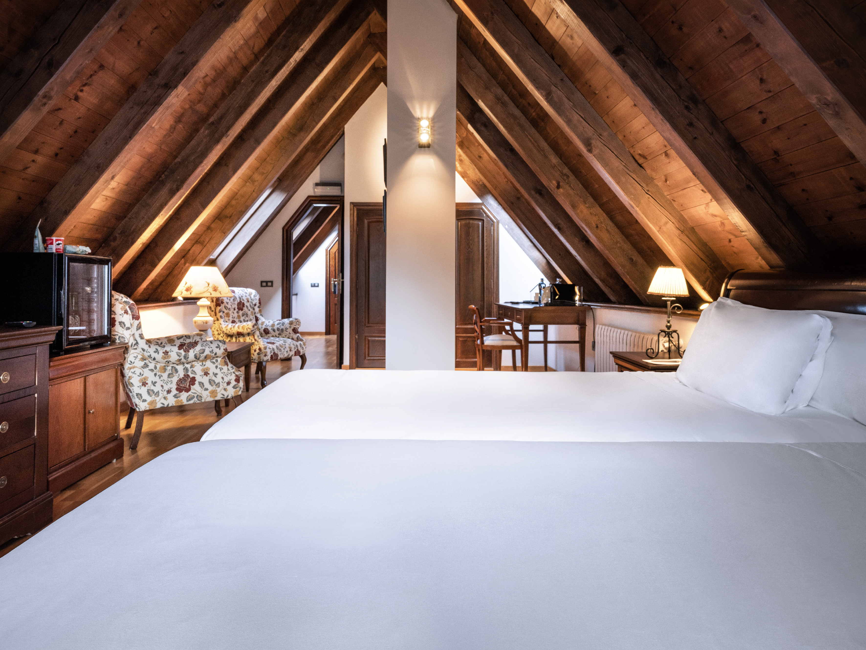 Cozy attic hotel room with exposed wooden beams, white beds, and inviting armchairs.