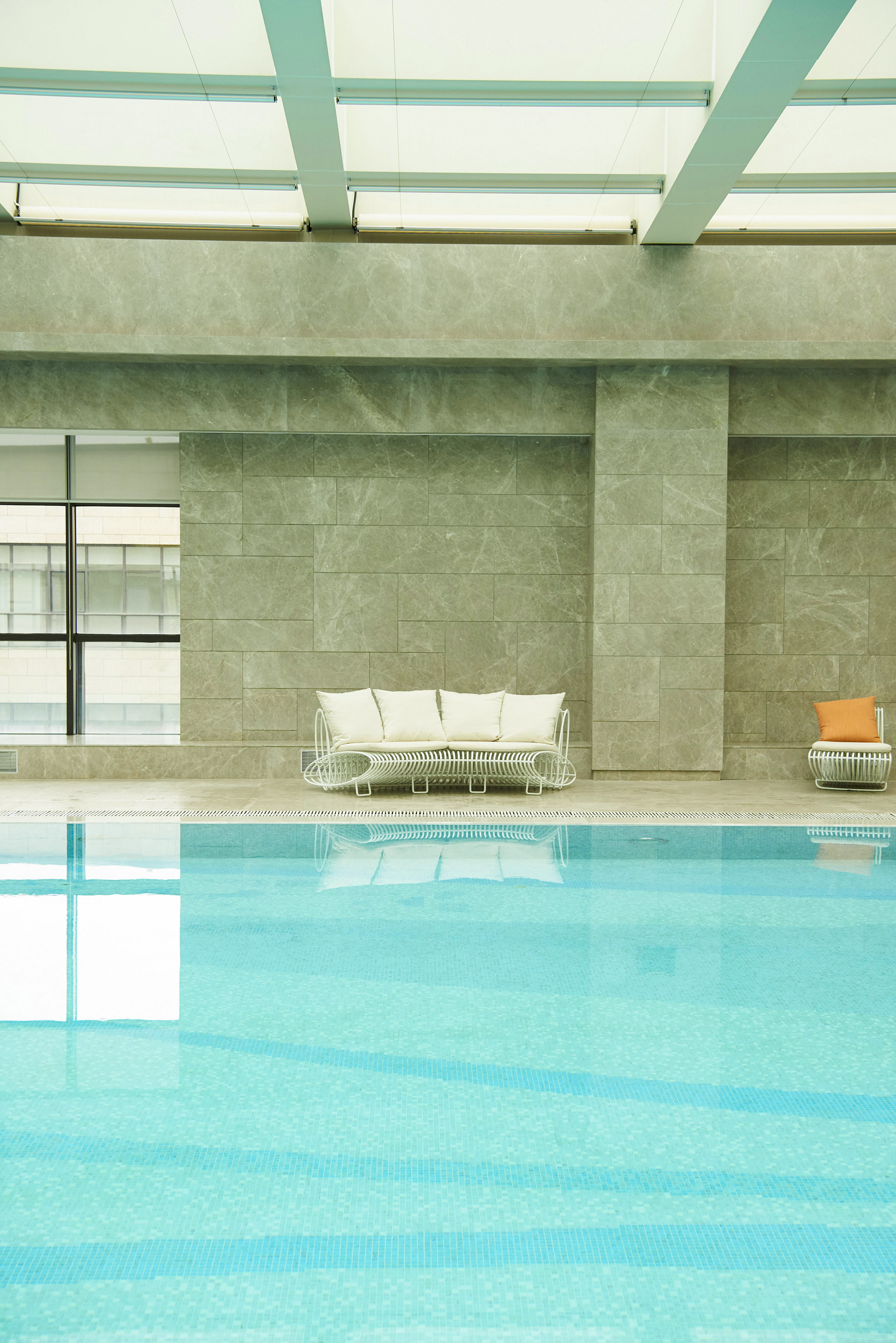 Modern indoor pool with white lounge sofa, blue tiled water, and marble walls.