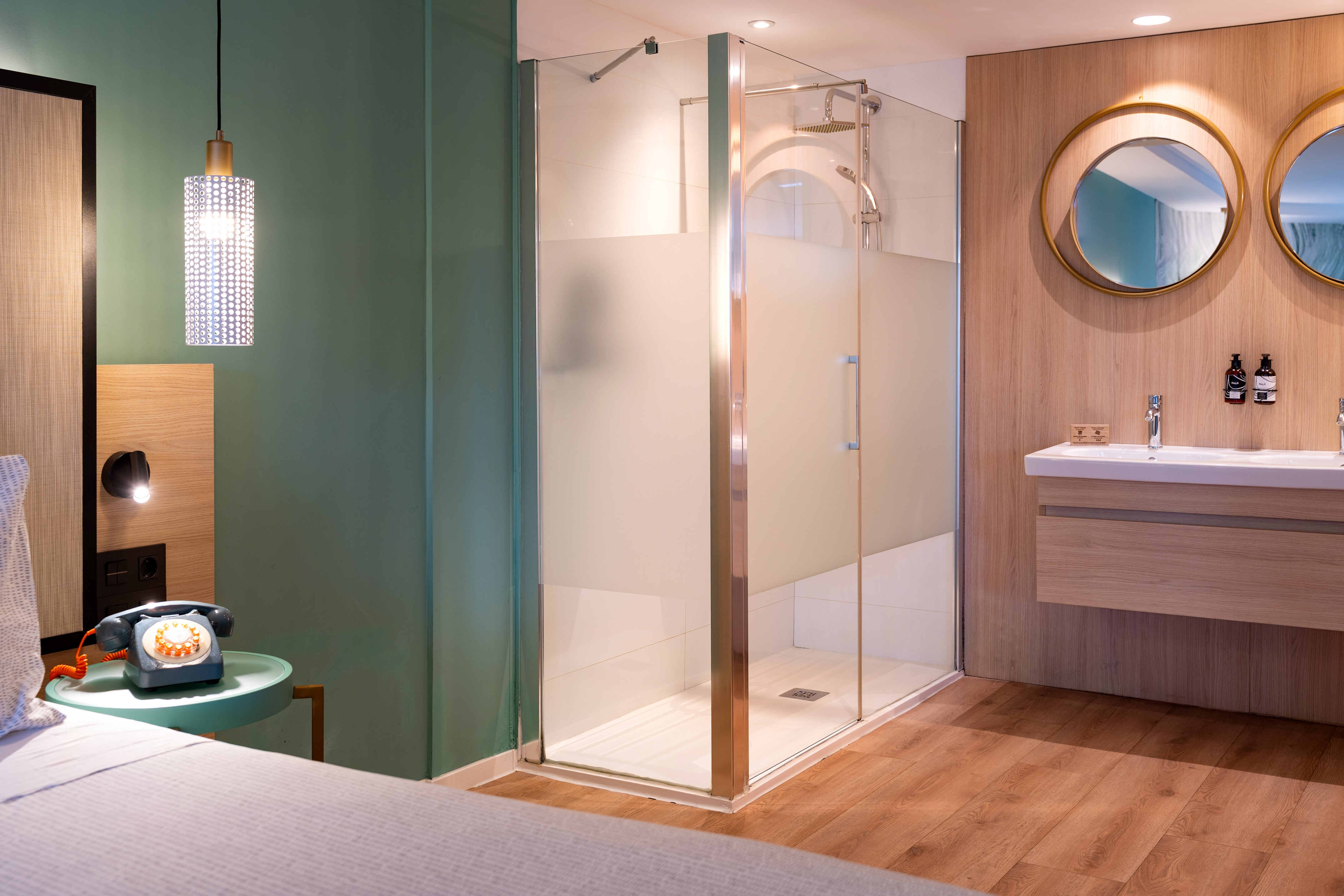 Modern hotel room: bed, vintage phone, glass shower, double vanity, wood floor.