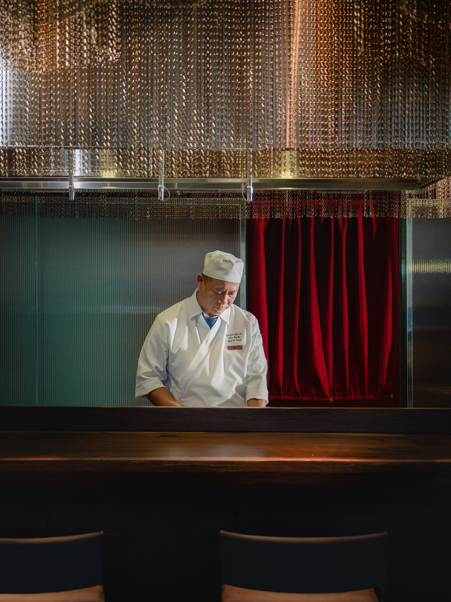 a man in a chef's hat standing in a room with chairs and a red curtain