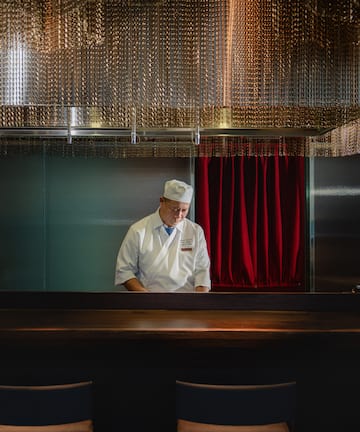 a man in a chef's hat standing in a room with chairs and a red curtain