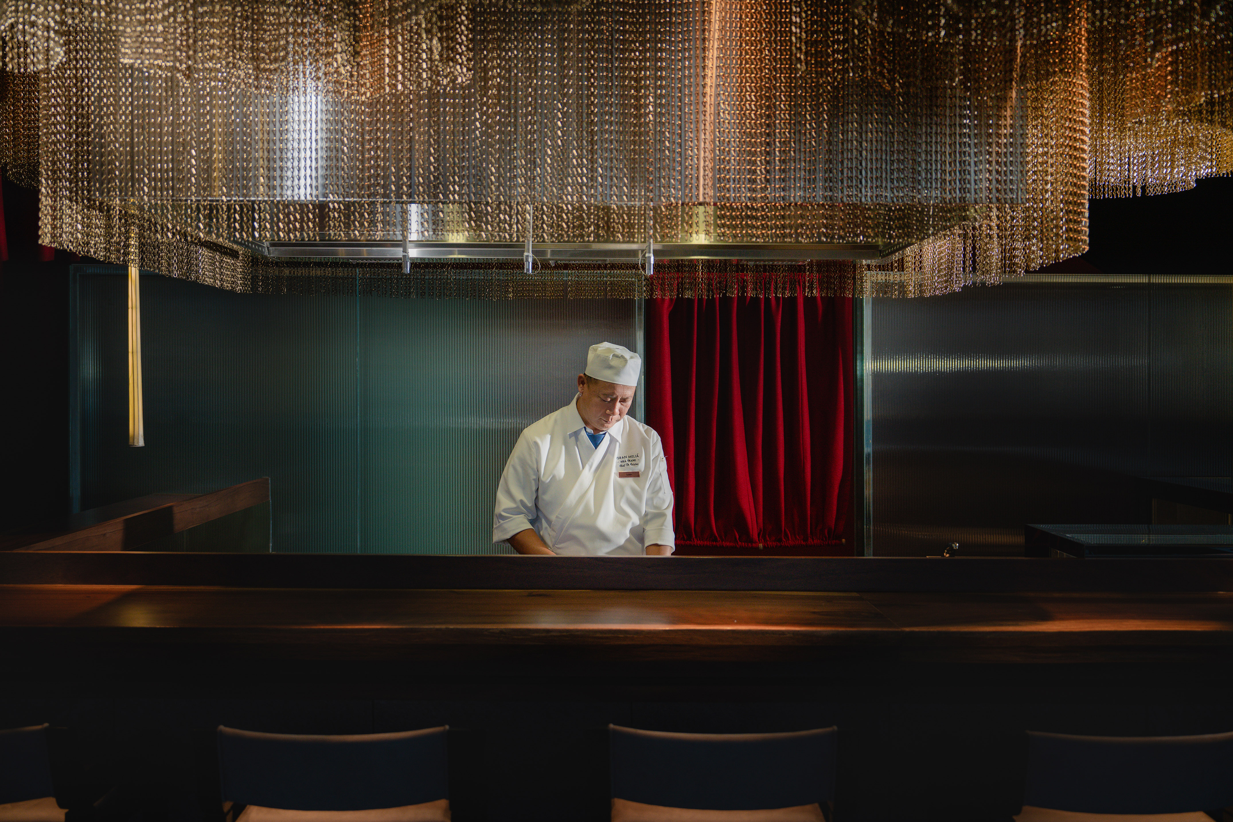 a man in a chef's hat standing in a room with chairs and a red curtain