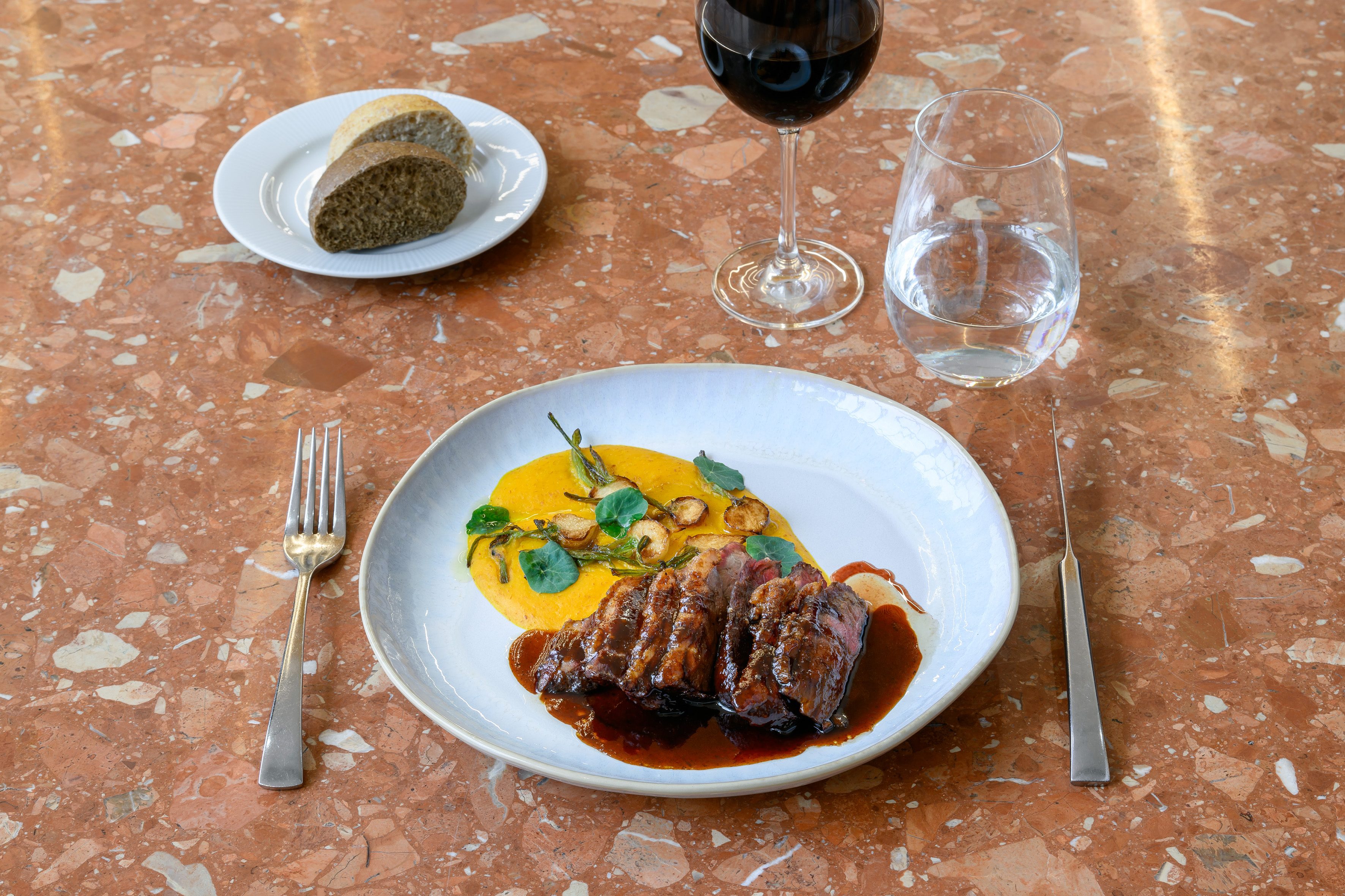 Gourmet sliced steak, yellow puree, red wine, bread on a terrazzo table.