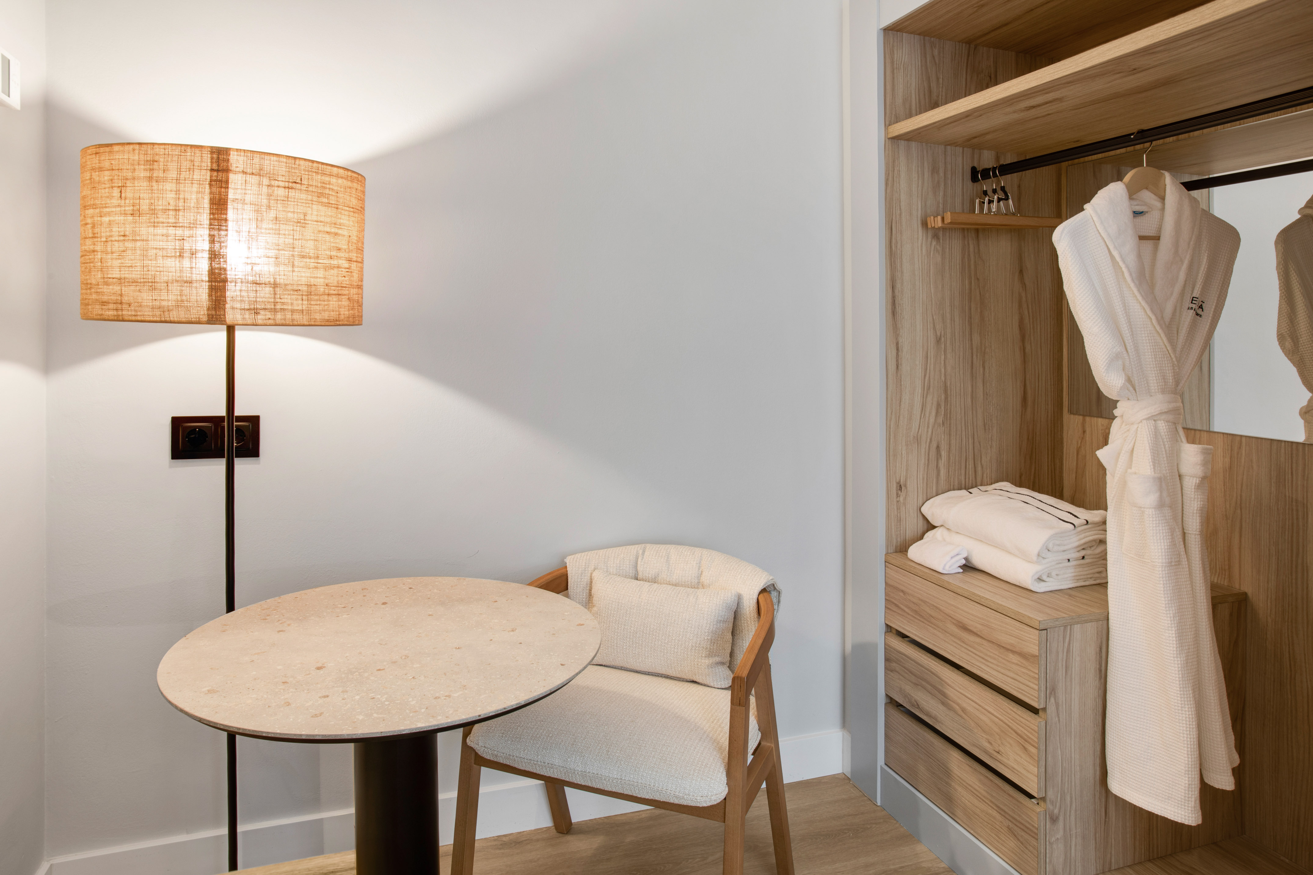 Hotel room corner with floor lamp, round table, chair, and open wooden wardrobe.