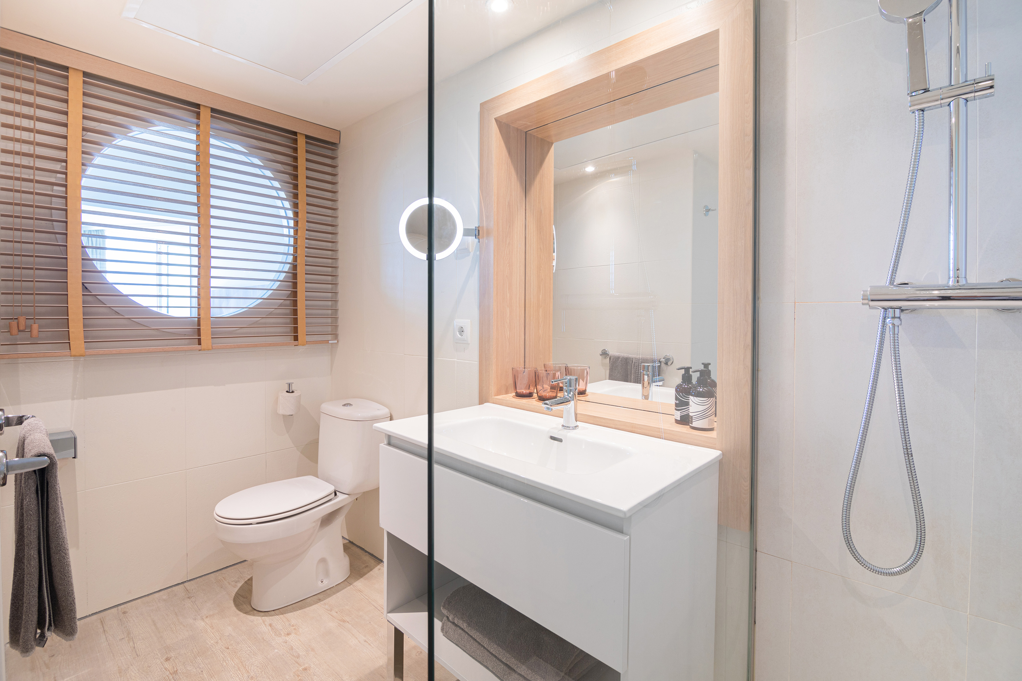 Modern bathroom with vanity, shower, toilet, and round window with wooden blinds.