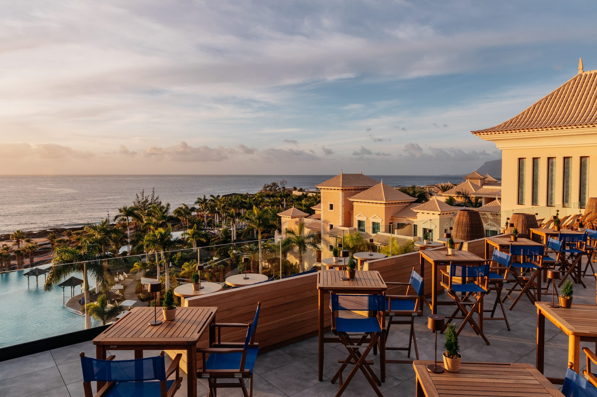 Rooftop restaurant terrace at sunset overlooking ocean, resort, and palm trees.