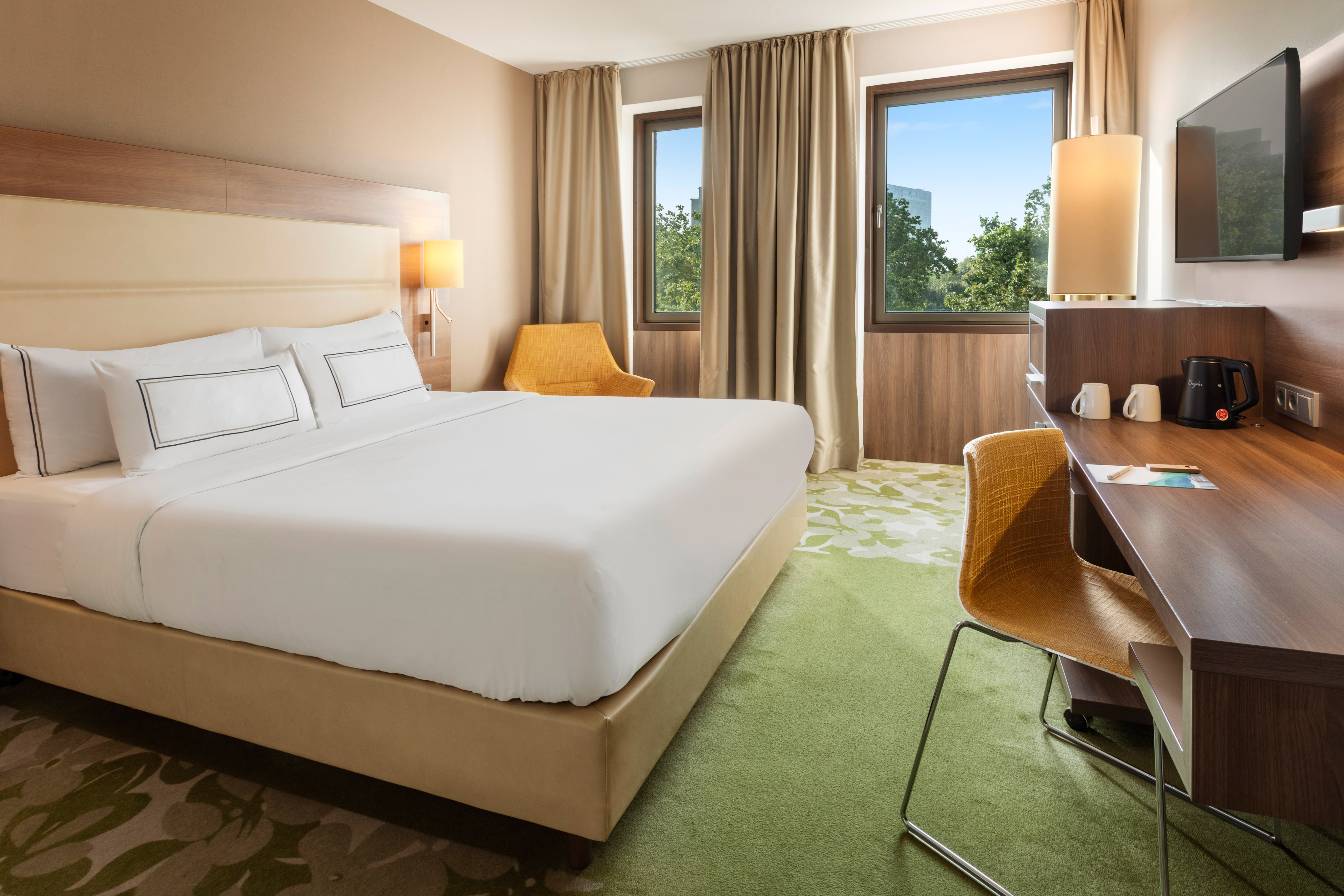 Modern hotel room with king bed, desk, armchair, and bright window view.