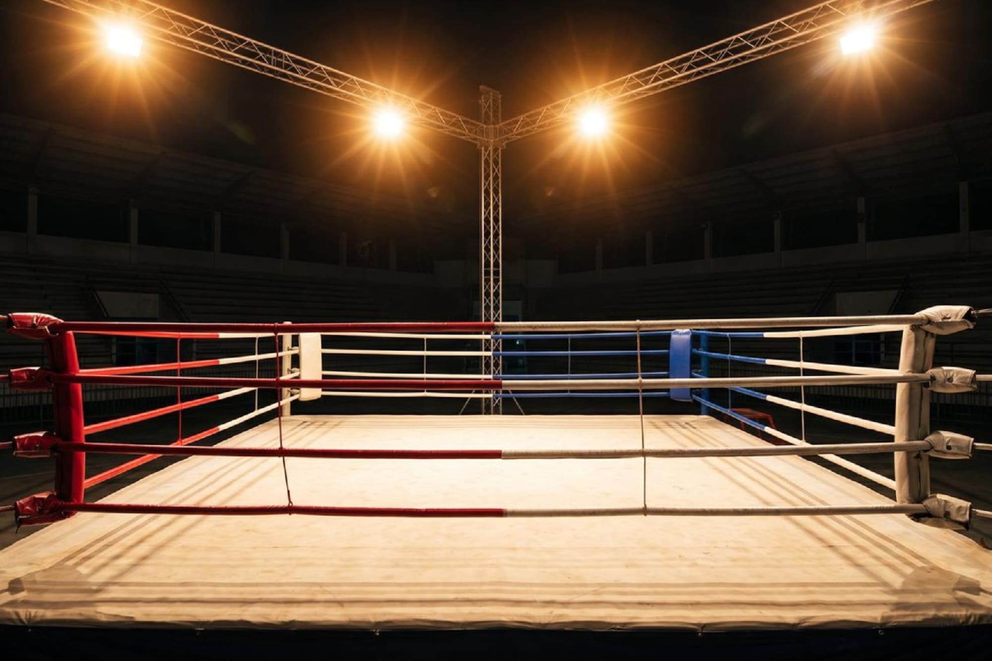 Empty boxing ring with red, white, blue ropes under bright arena spotlights.