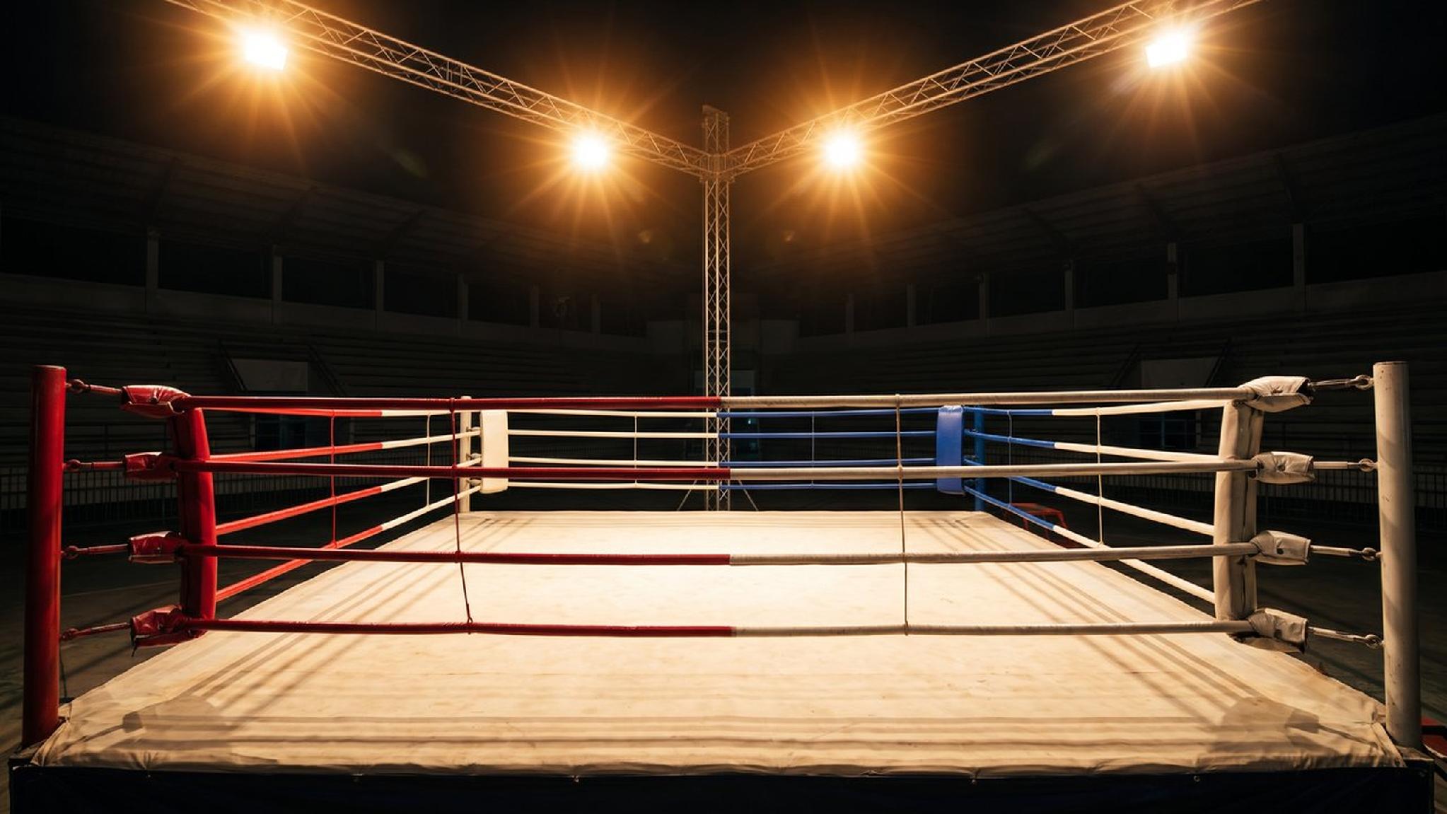 Empty boxing ring with red, white, blue ropes under bright arena spotlights.
