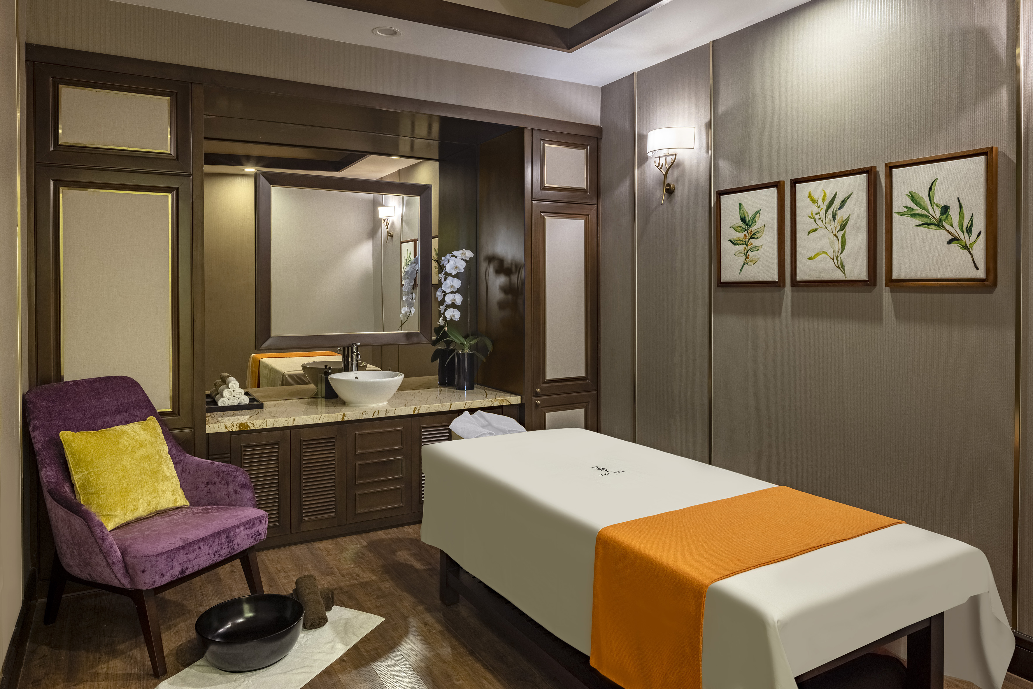 Luxury spa room: massage table, sink vanity, plush armchair, botanical art.