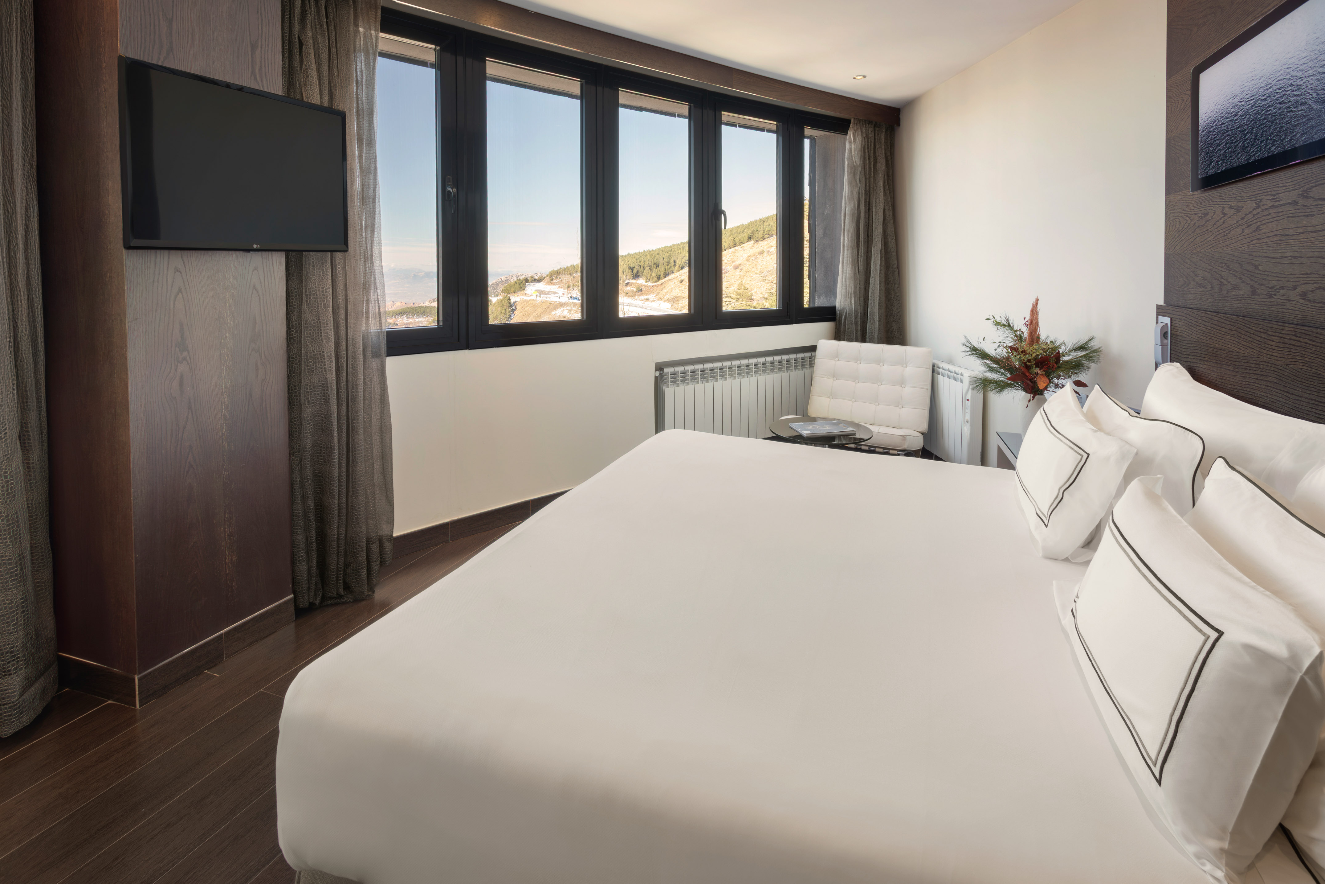 Modern hotel bedroom with white bed, TV, and winter mountain view.