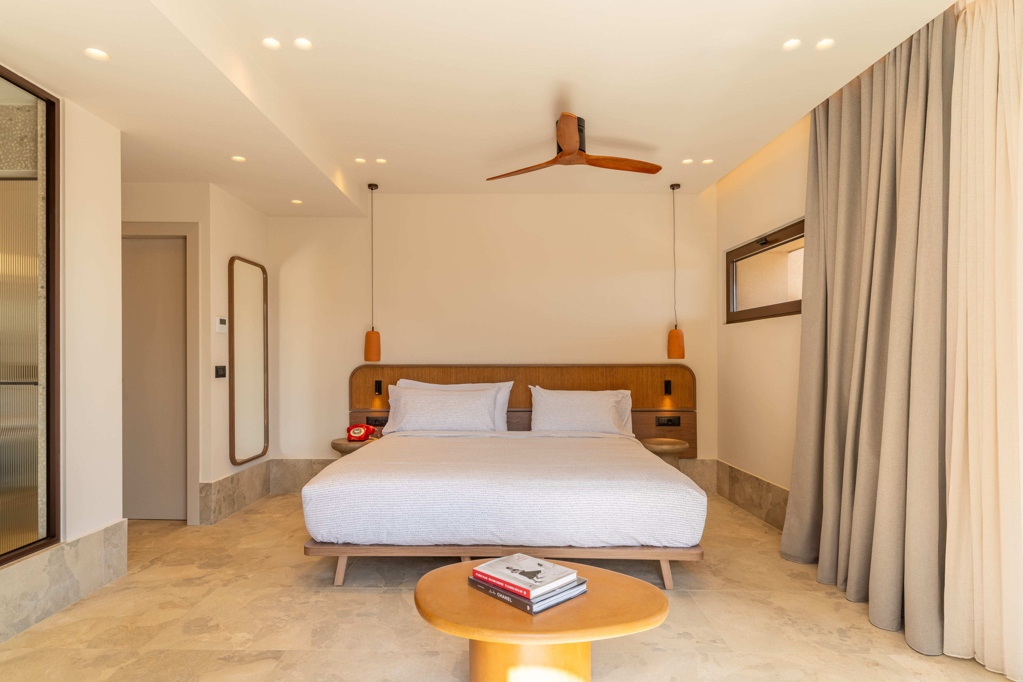 Contemporary hotel bedroom with wooden bed, ceiling fan, neutral tones, and tiled floor.