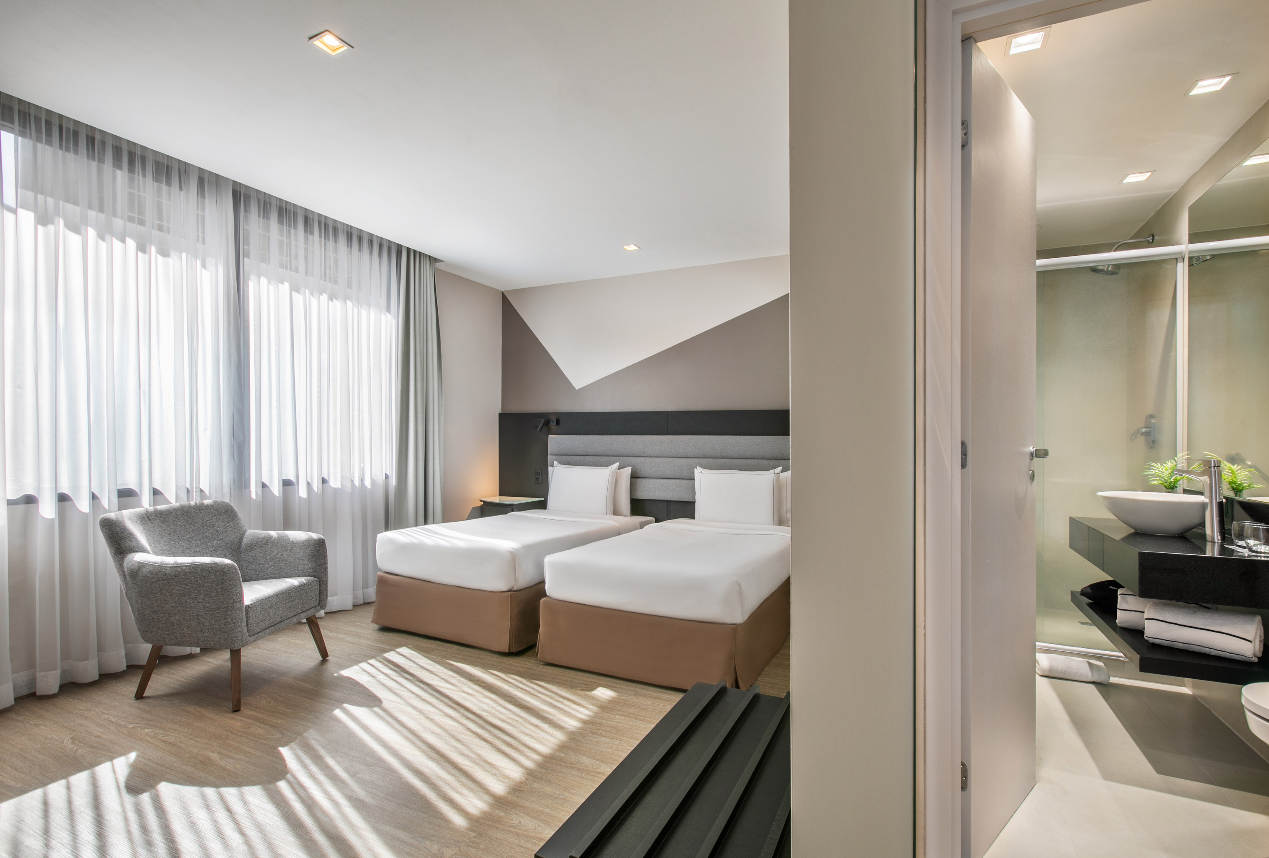 Modern hotel room with twin beds, armchair, and open view to bathroom.