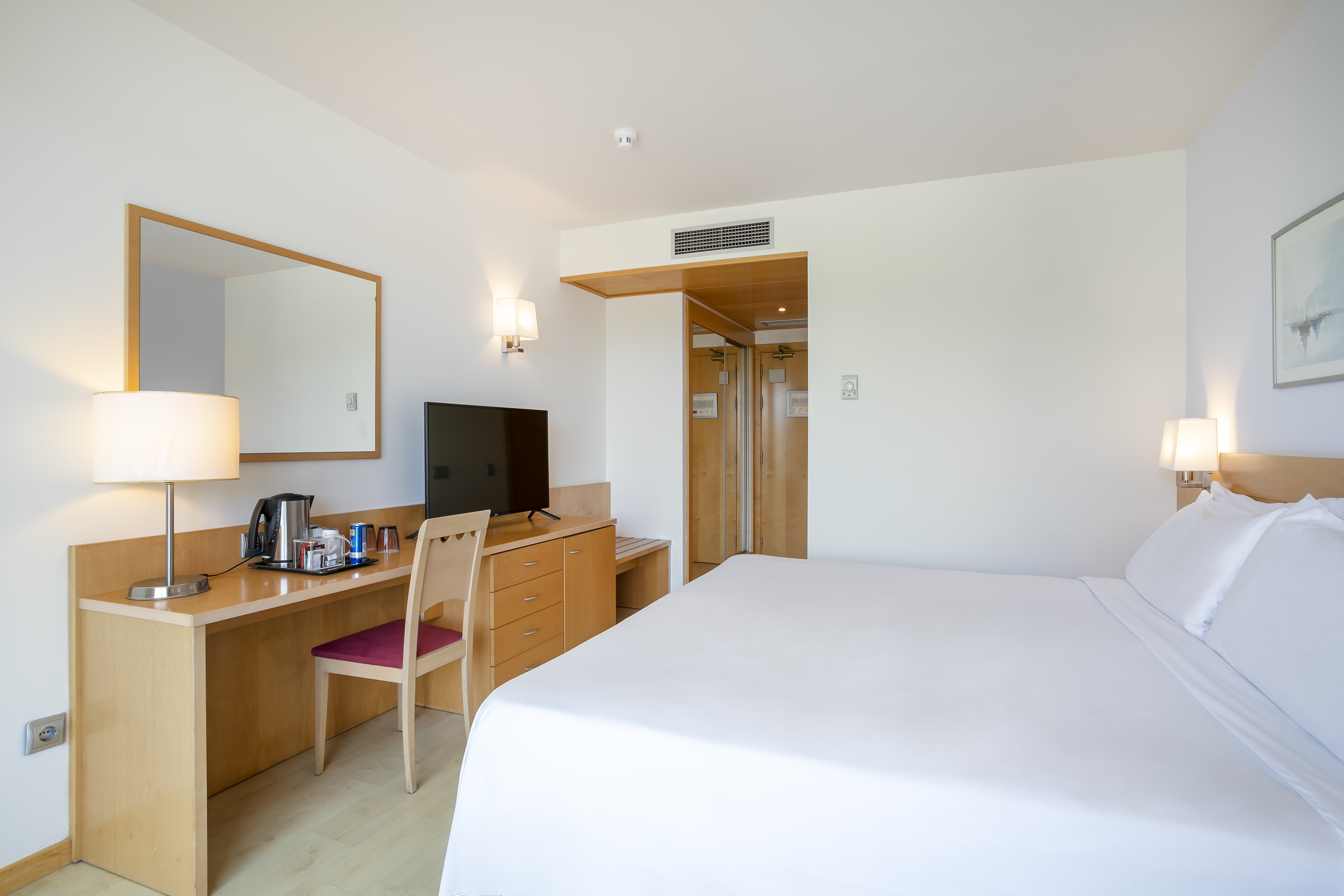 Bright hotel room with white bed, wooden desk, TV, and a kettle.