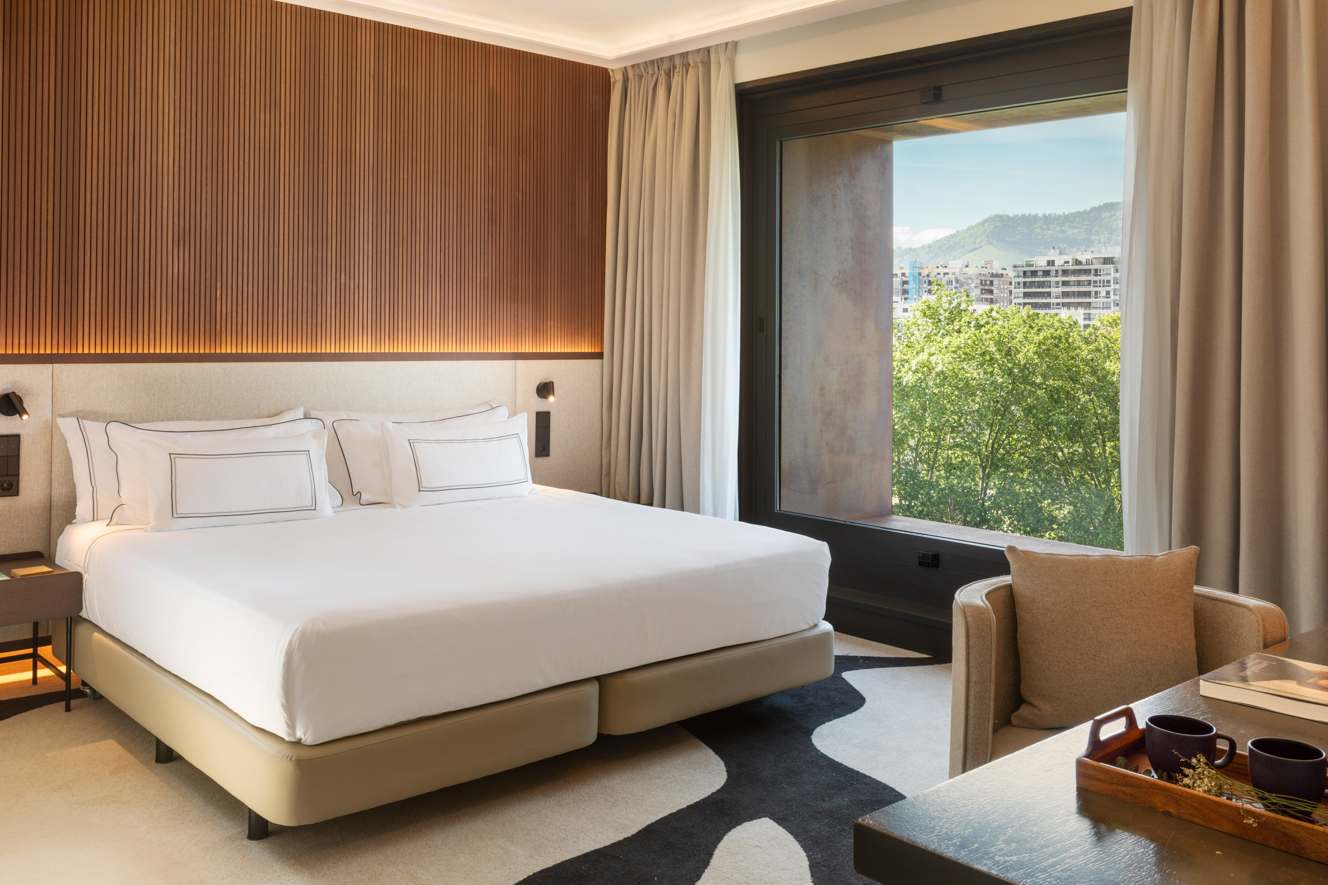 Modern hotel bedroom with bed, wood-paneled wall, and city view from window.