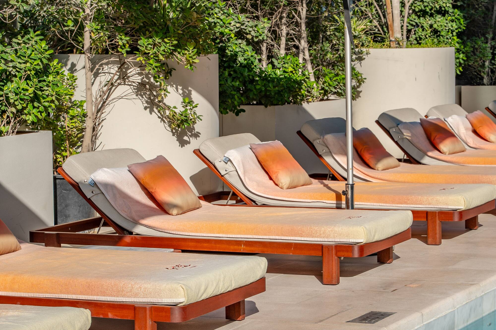 Poolside with sun loungers, orange pillows, and white umbrellas amidst green plants.