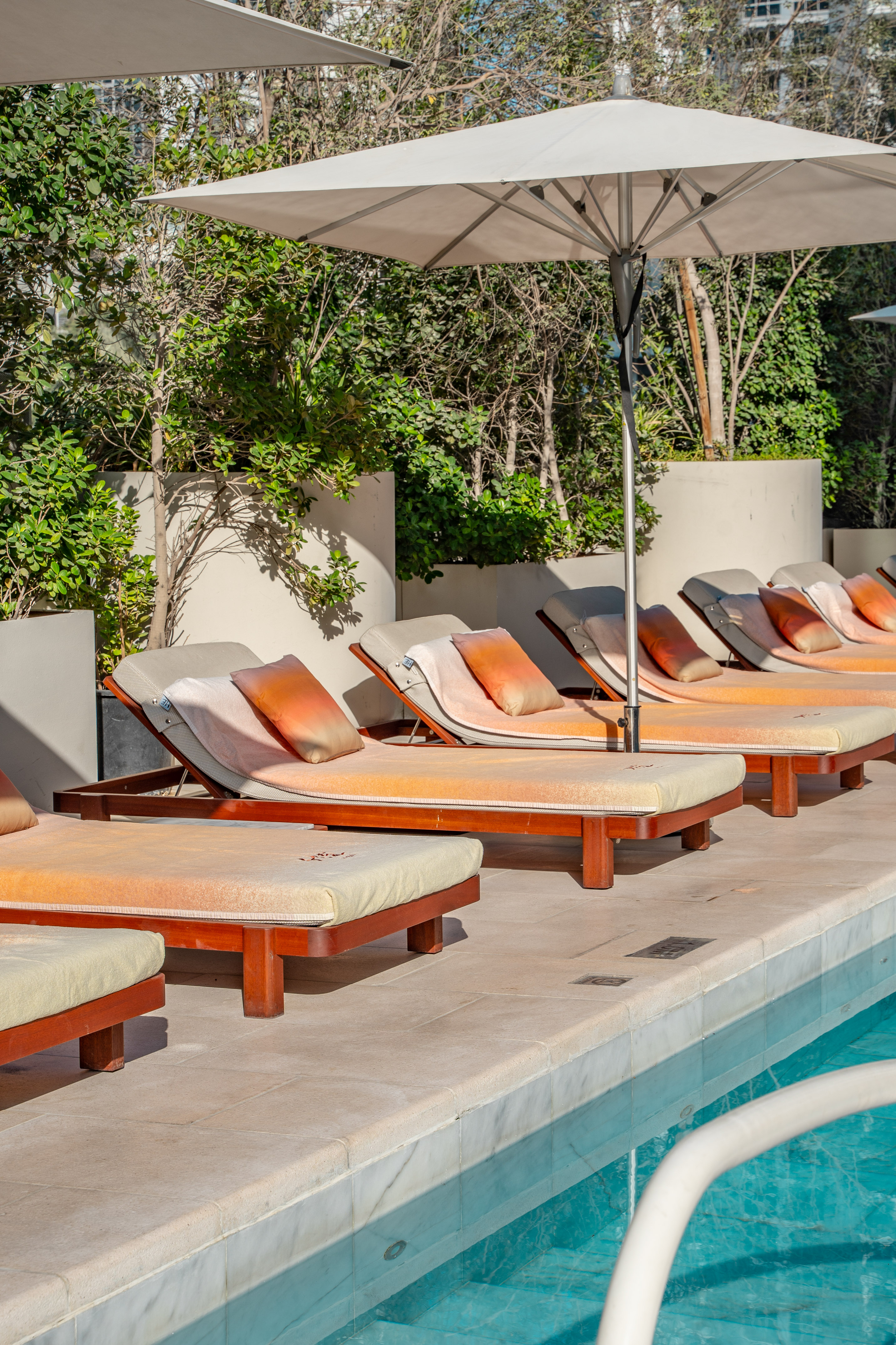 Poolside with sun loungers, orange pillows, and white umbrellas amidst green plants.