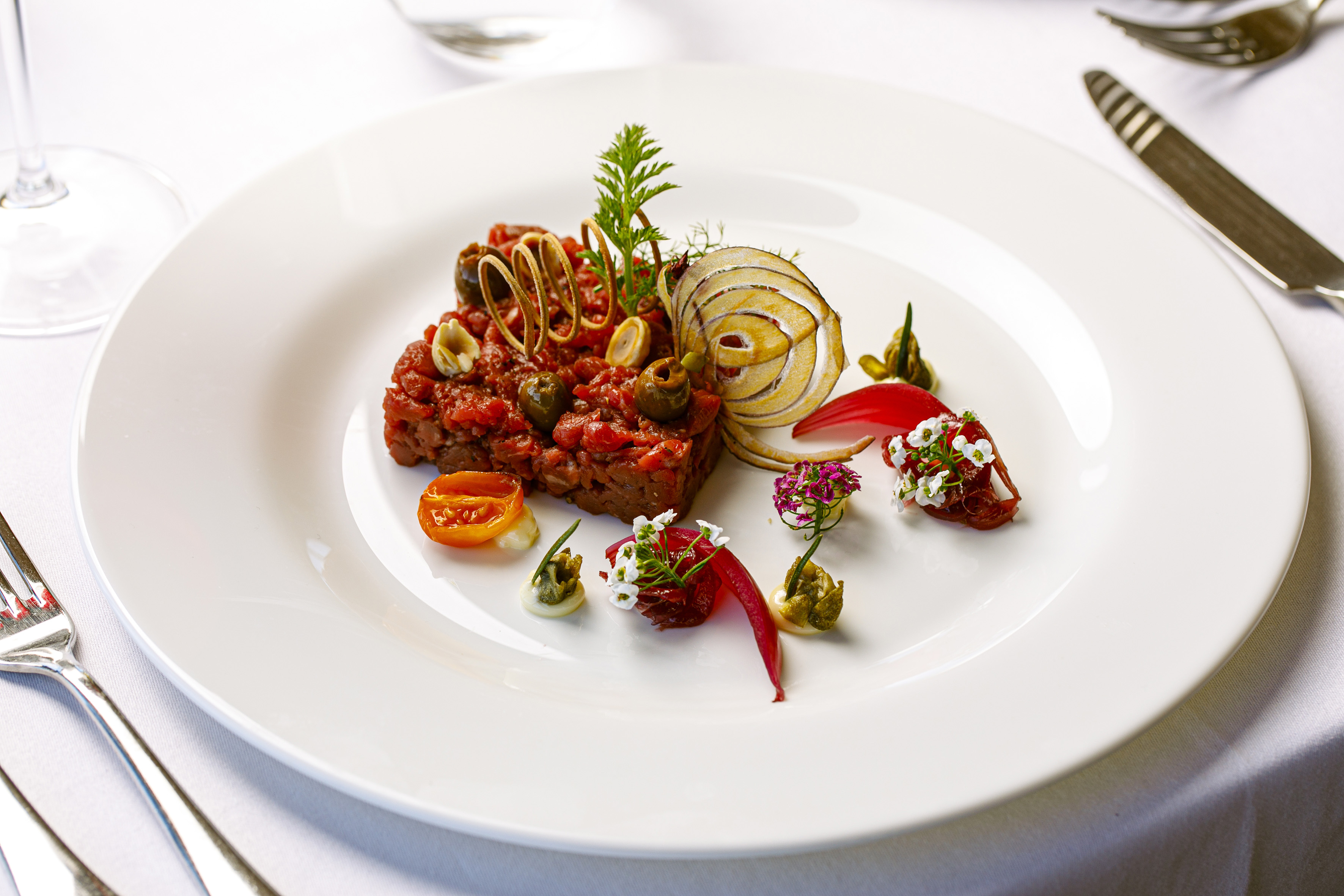 Gourmet tartare dish with olives, spiral garnish, and delicate edible flowers.