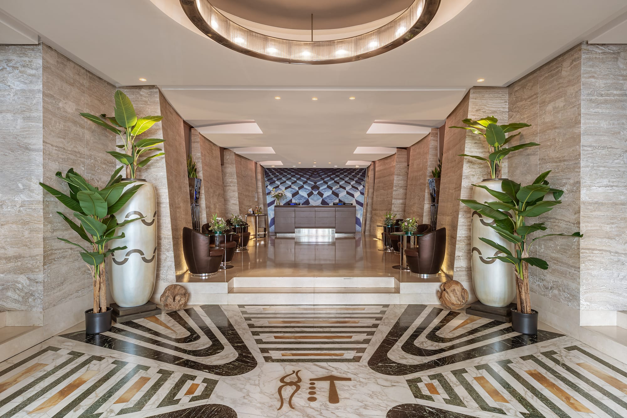 Luxury hotel lobby with ornate marble floor, tall plants, and reception area.