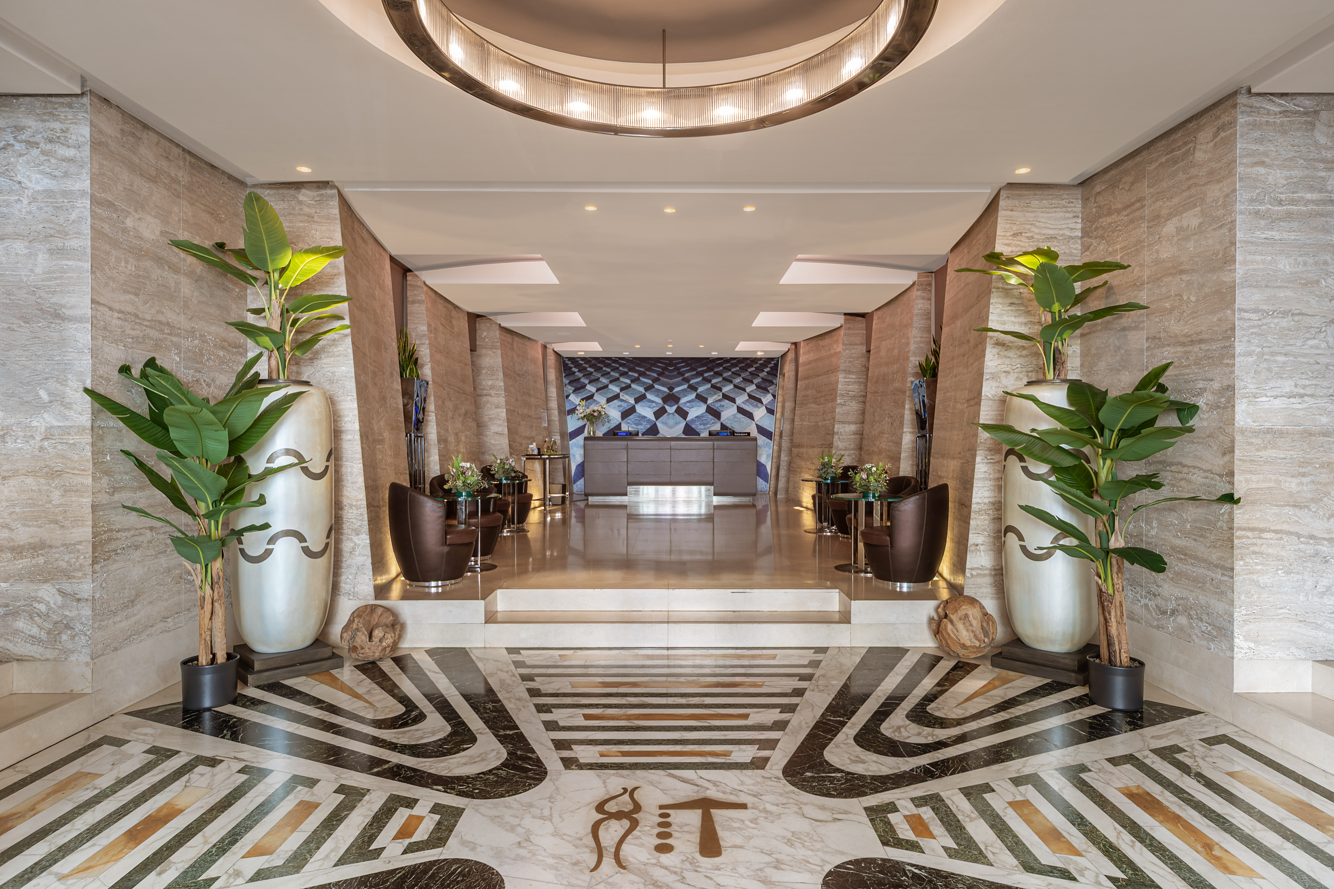 Luxury hotel lobby with ornate marble floor, tall plants, and reception area.